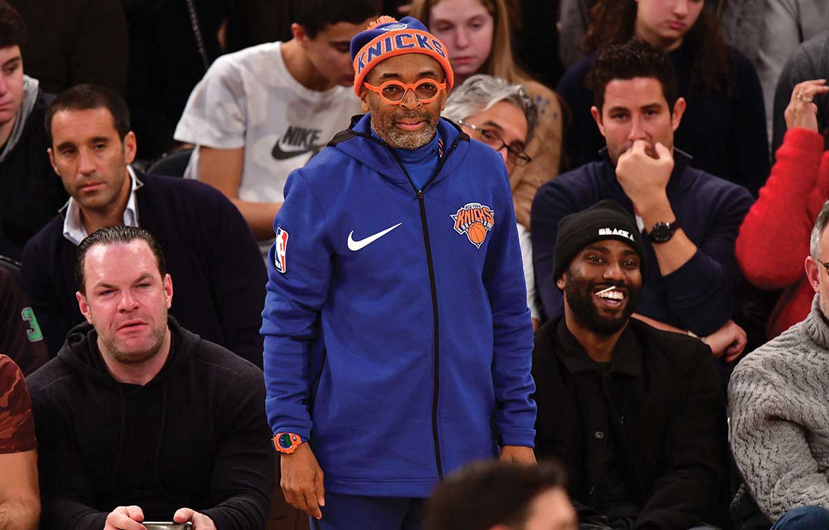 Spike Lee in einem Knicks-Outfit in einer Sporthalle neben mehreren Personen trägt am Handgelenk eine orange-lilane G-SHOCK Digitaluhr.
