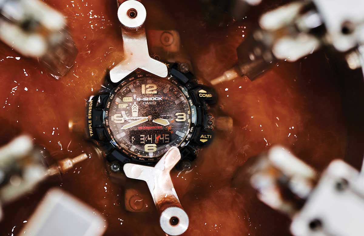 Eine G-SHOCK Armbanduhr mit analoger Anzeige und massivem Gehäuse ist in einer Maschine eingespannt, drum herum ist eine rötliche Flüssigkeit
