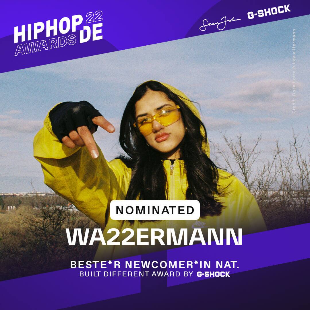 Rapperin Wa22ermann war als beste Newcomerin 2022 national nominiert, sie trägt eine gelbe Jacke, eine gelbe Brille und zeigt ein Peace-Zeichen in die Kamera.