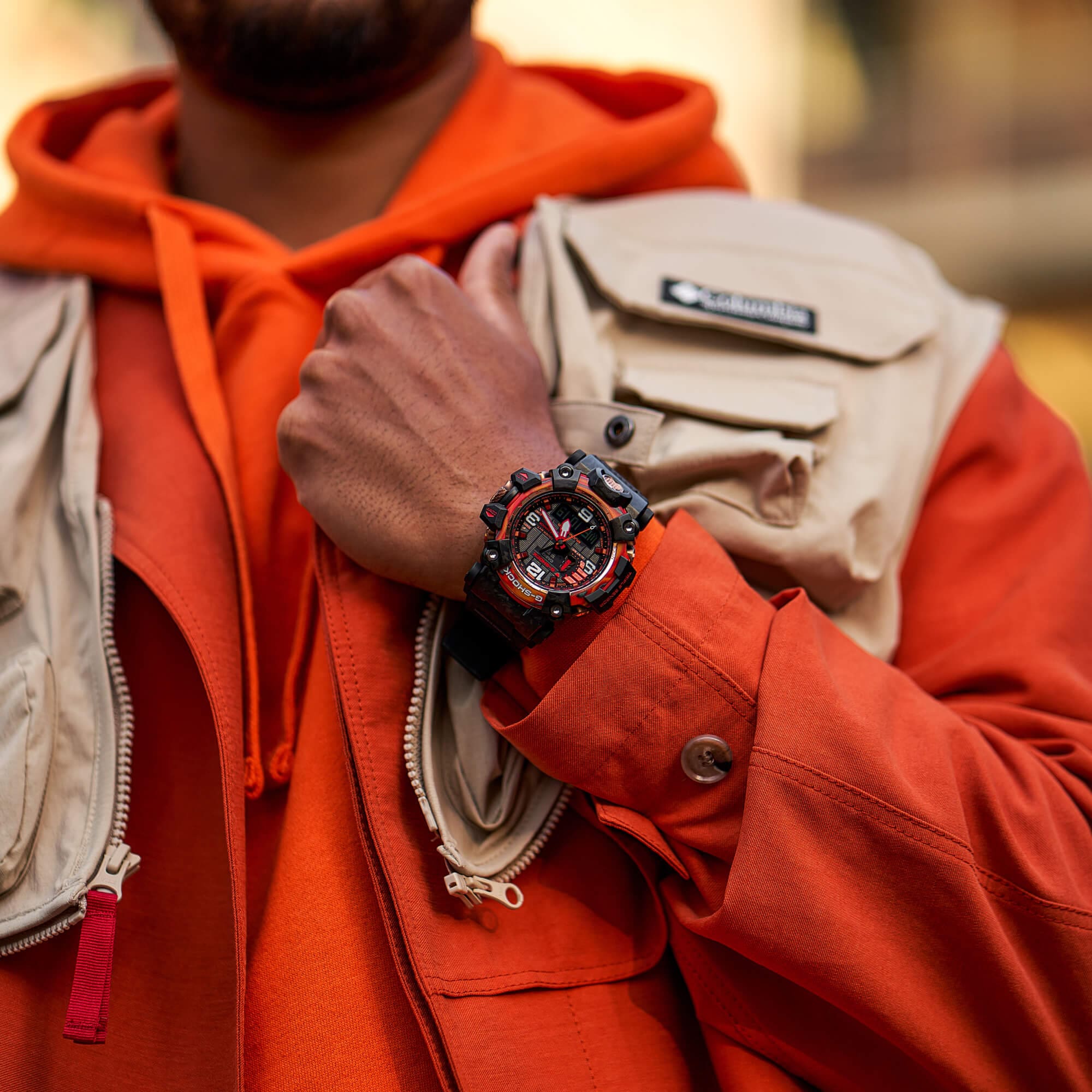 Ein Mann in orangenem Hoodie und beiger Weste trägt die GWG-2040FR-1AER Armbanduhr von G-SHOCK am Handgelenk.