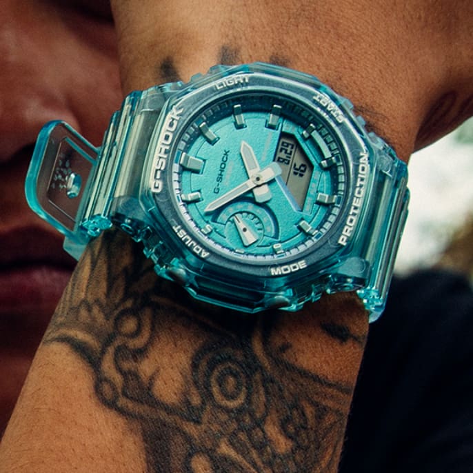 Eine Person hält sie die türkis-durchsichtige Armbanduhr "GMA-S2100SK-2A" vor ihr Gesicht. Das Gehäuse der Uhr ist achteckig, die Anzeige rund und analog.