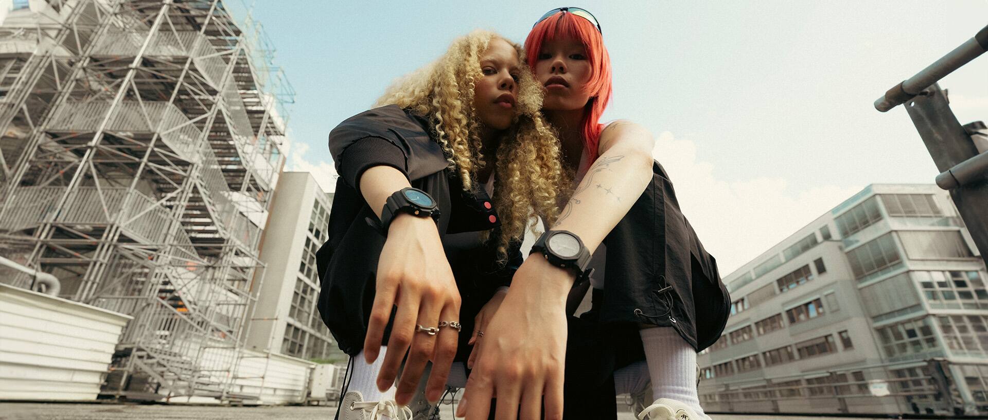 Zwei junge Frauen sitzen in der Hocke und schauen herunter auf die Kamera. Sie tragen dunkle Kleiding, helle Sneaker und schwarze G-SHOCK Analoguhren am Handgelenk.