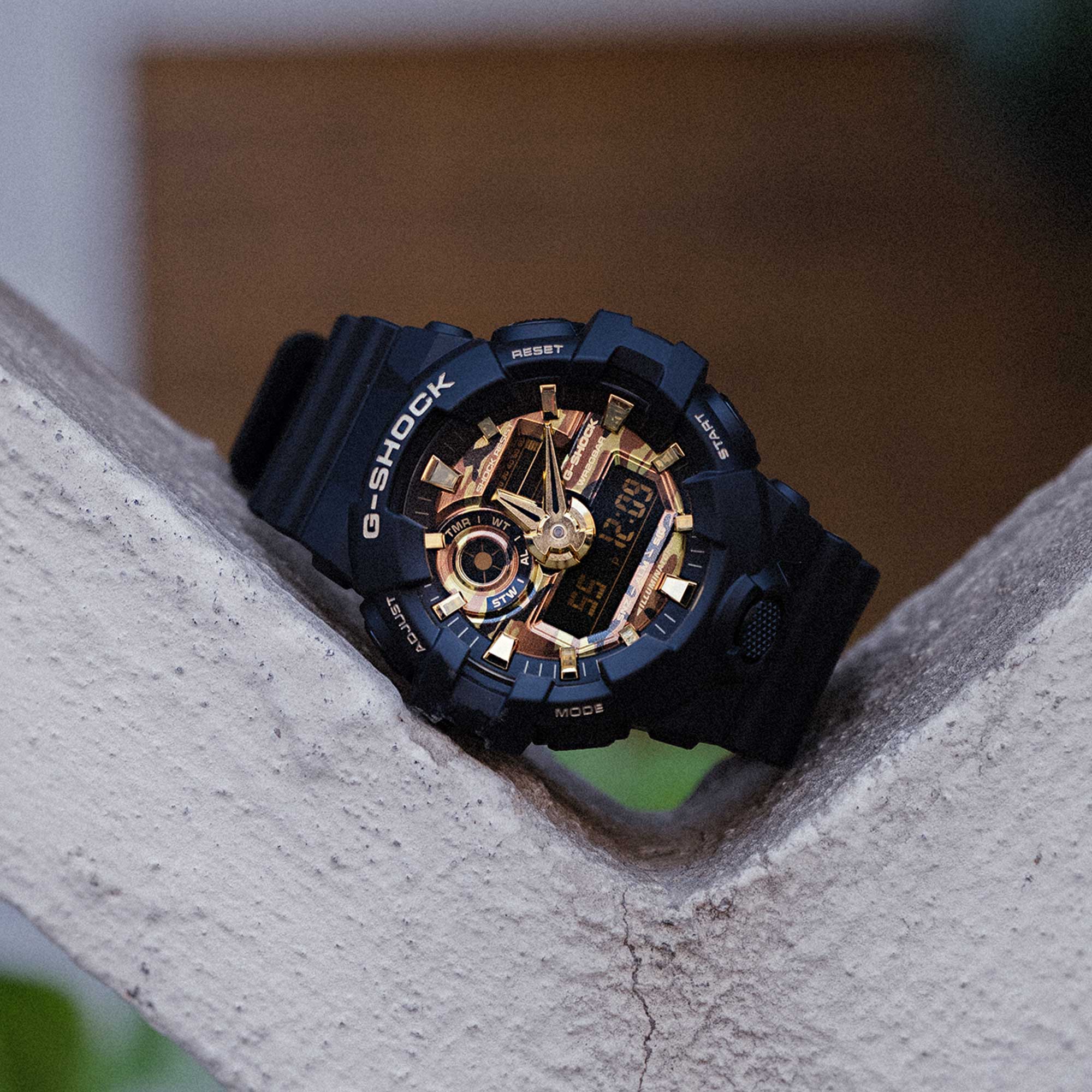 Die schwarze G-SHOCK GA-700CF-1A mit goldfarbenem Zifferblatt liegt schräg auf einem hellen Ast vor einem unscharfen Hintergrund in Braun- und Grüntönen.