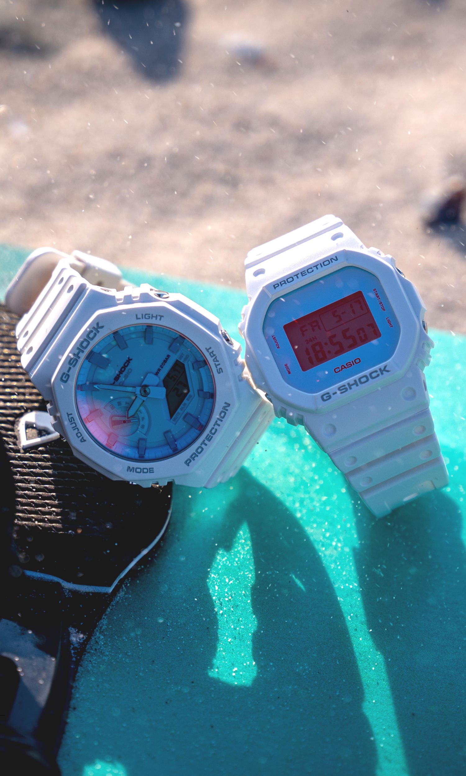 Die Armbanduhren GA-2100TL-7A und DW-5600TL-7 mit weißem Armband auf einem Surfboard im Sand