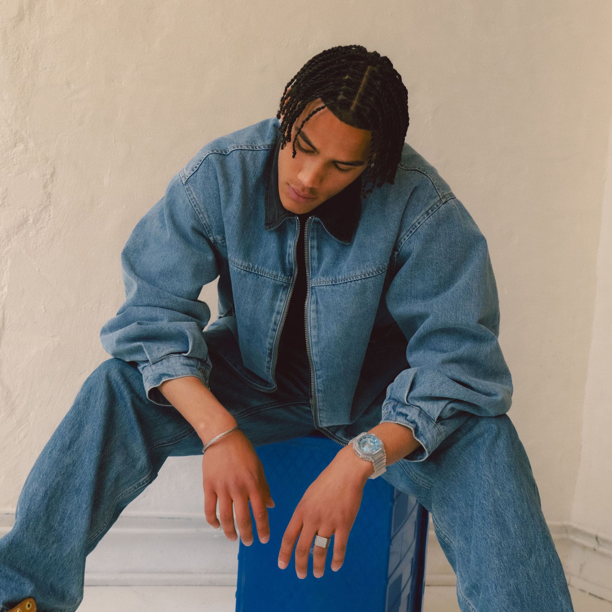 Ein Mann in Jeanskleidung sitzt auf einem blauen Hocker und beugt sich nach vorne, am Handgelenk trägt der die G-SHOCK GA-2100BM mit hellblauem Zifferblatt.