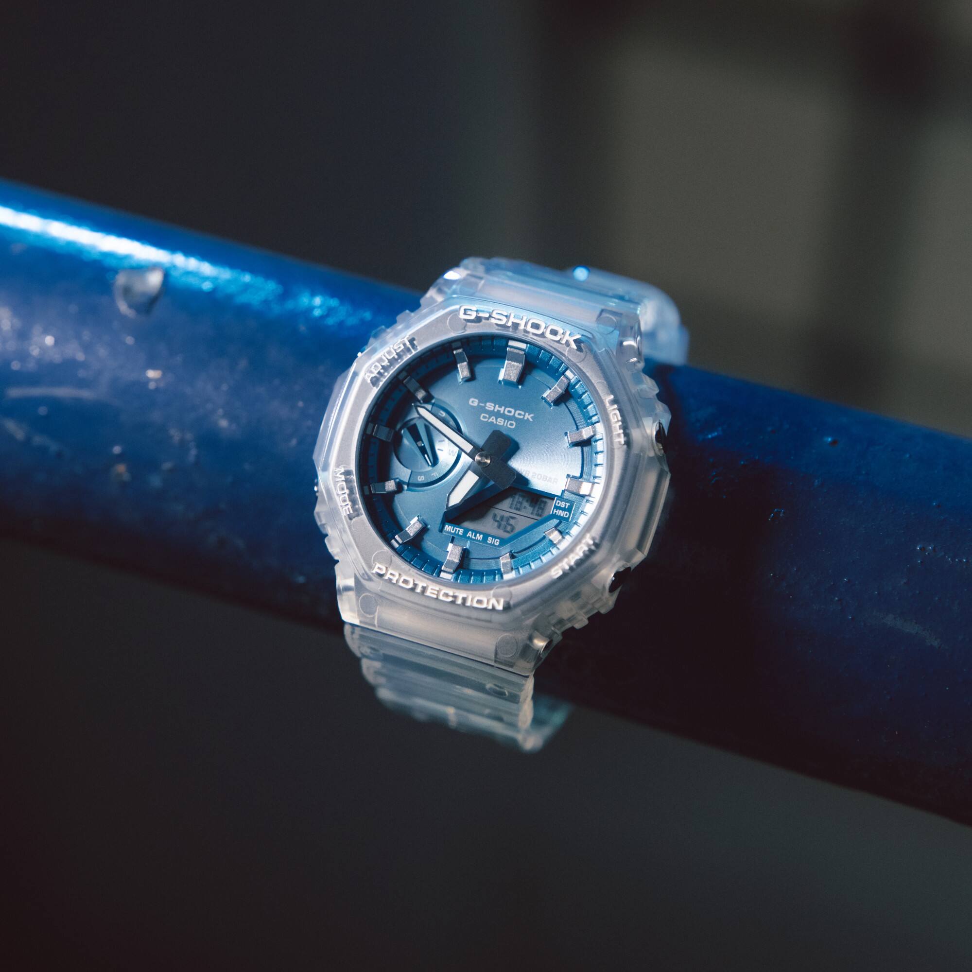 Die GA-2100BM Armbanduhr von G-SHOCK mit transparentem Armband und achteckigem Gehäuse sowie hellblauem Zifferblatt an einer blauen Stange hängend.