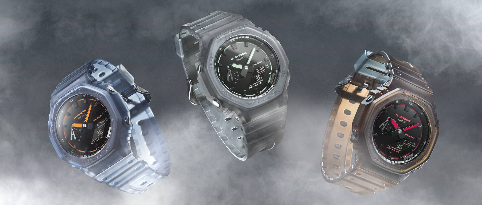 Drei verschiedenfarbige G-Shock GA-2100K Armbanduhren mit analog-digitalen, achteckigen Anzeigen und transparenten Armbändern schweben nebeneinander im Nebel.