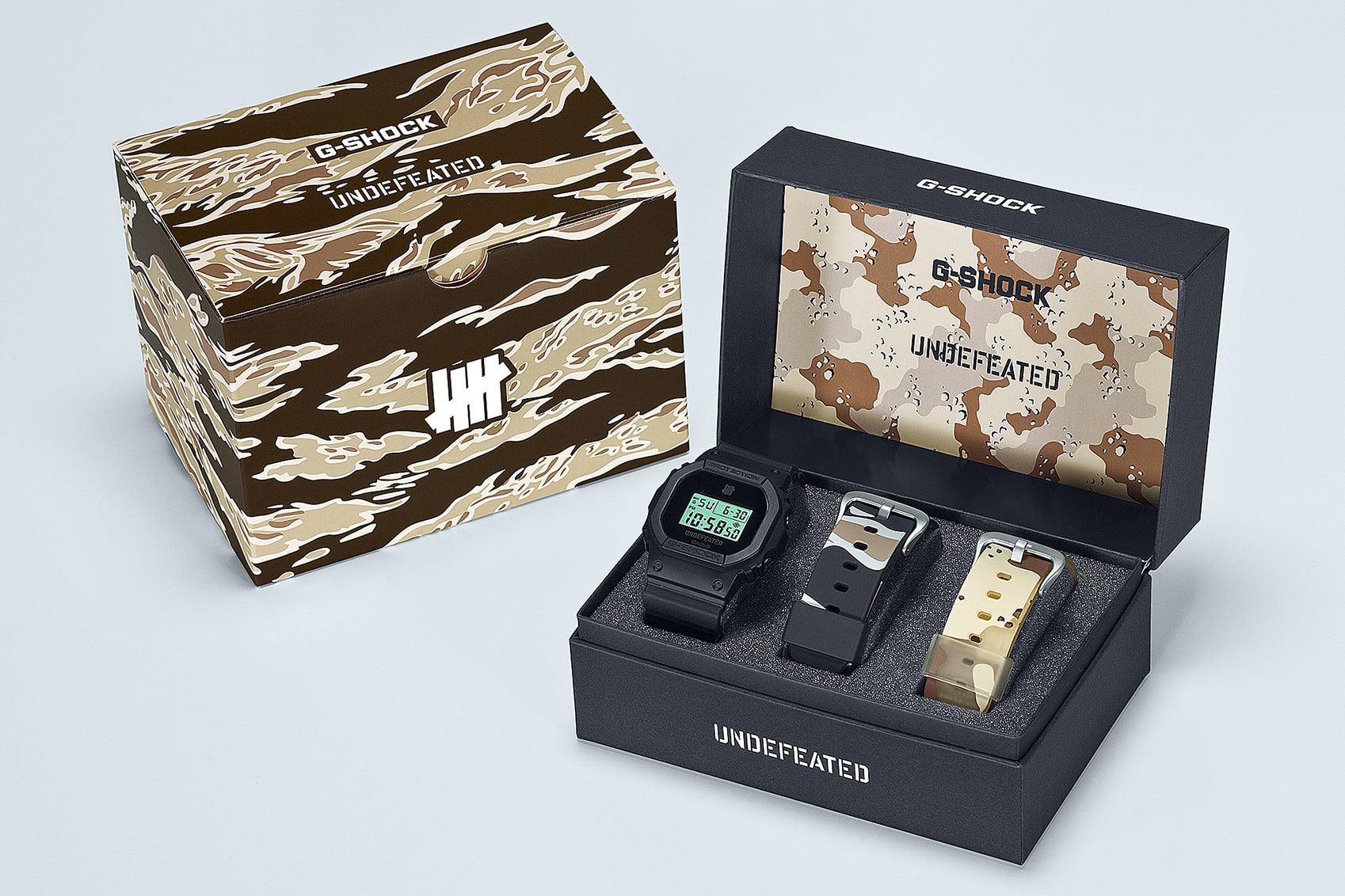 Neben der offenen Verpackungsbox der DWE-5600UD-1 mit der Uhr und ihren beiden Wechselarmbändern steht die Umverpackung in Camouflage-Design
