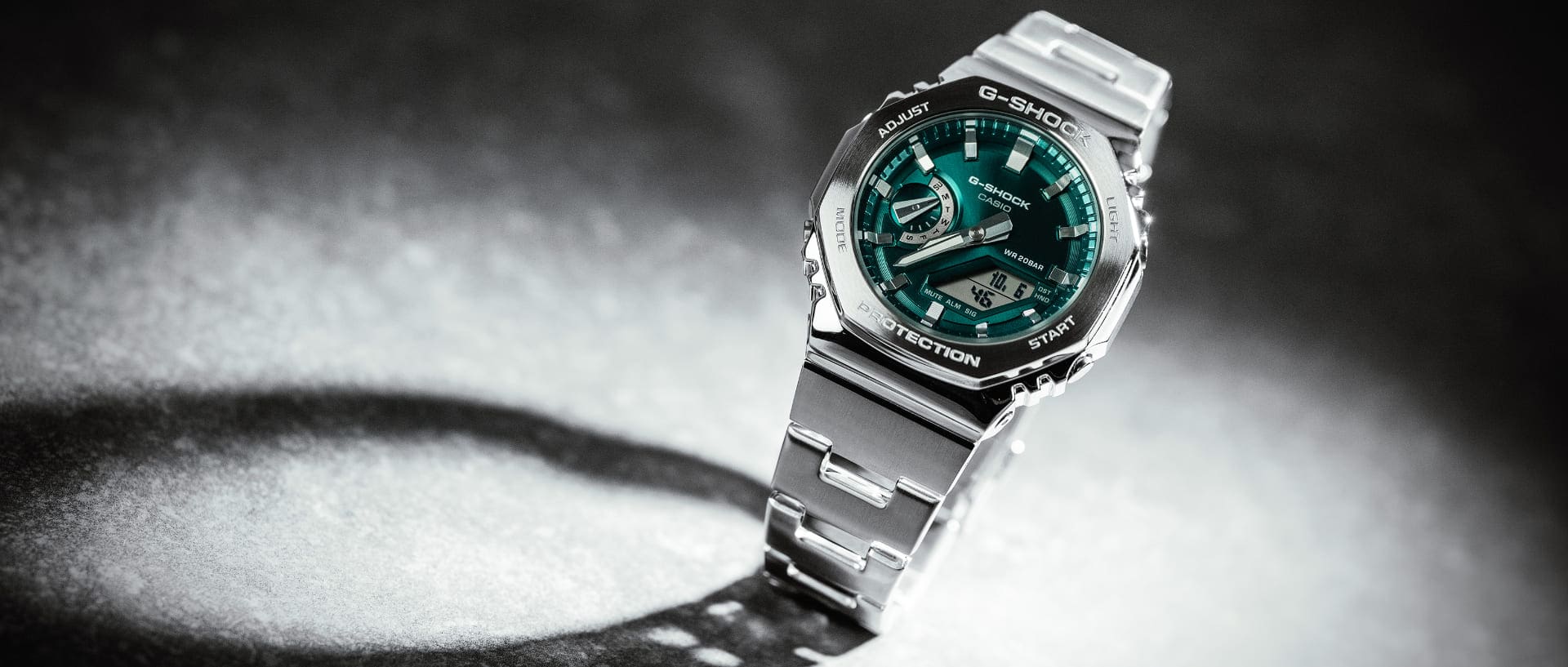 Die silberfarbene G-SHOCK GM-2110D mit Gliederarmband aus Stahl, grün-blauem, analogem Zifferblatt auf einem weiß-grauem Untergrund.