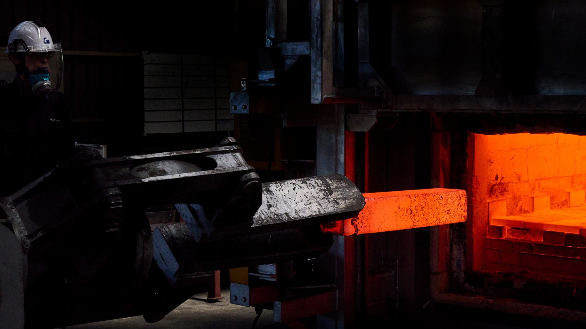 Arbeiter mit Helm neben heißem Metallstück;Glühendes Metall wird in Ofen geschoben;Dunkle, industrielle Atmosphäre mit intensivem orangenem Licht.