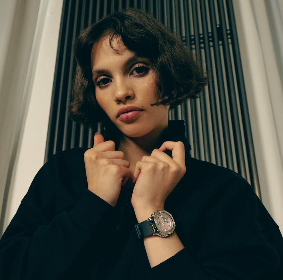 Eine Frau hält den Kragen ihres Pullovers fest. Ihr Handgelenk ziert die Armbanduhr GM-S2110-4AER von G-SHOCK mit Metalllünette und rosafarbenem Zifferblatt