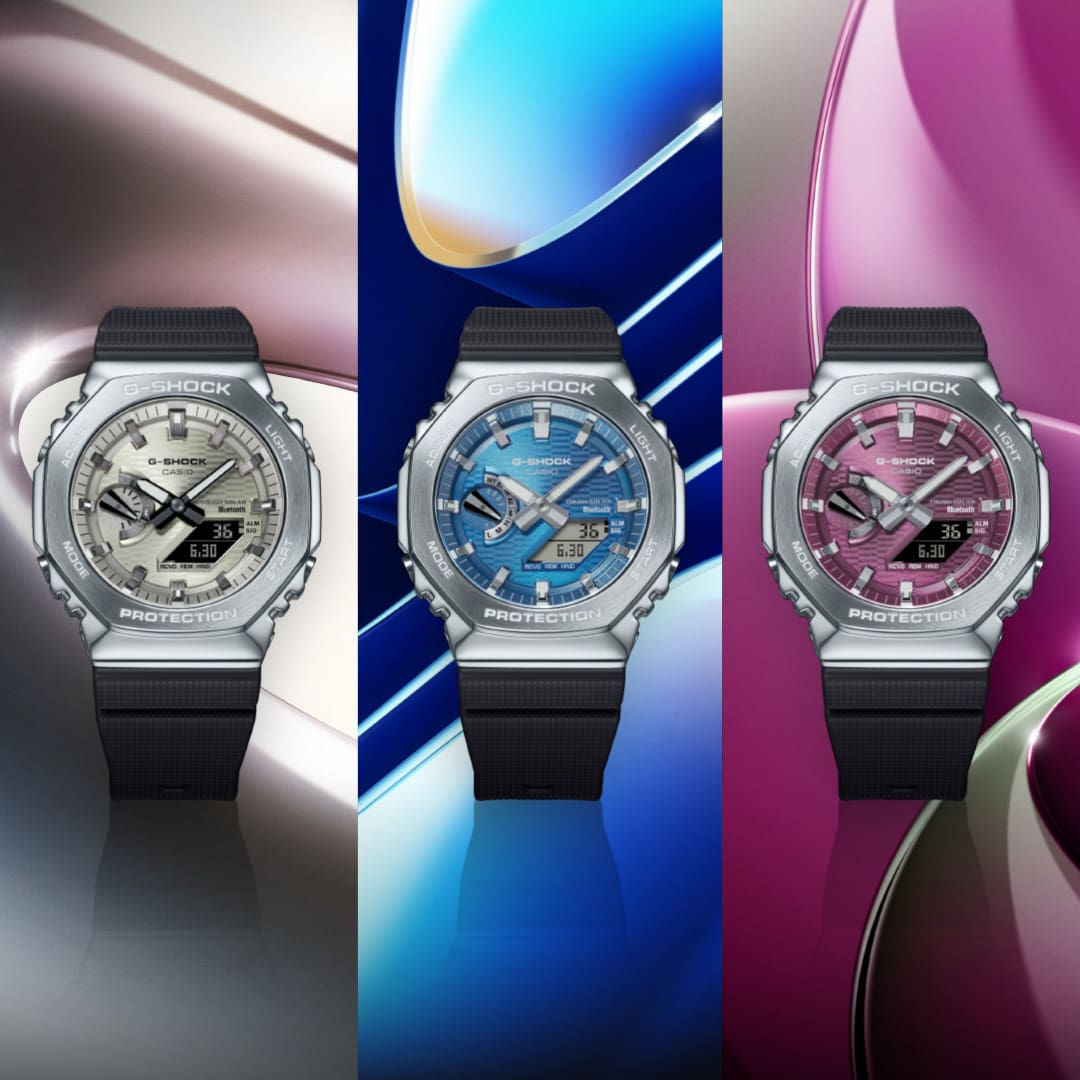 Drei Armbanduhren der GBM-2100-Serie mit schwarzem Resinband und Metalllünette sowie analogen Zifferblättern in shimmerndem grau-weiß, hellblau und pink.