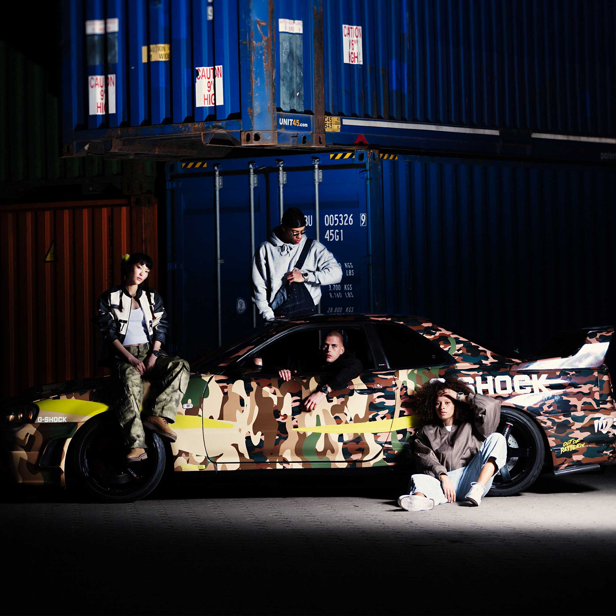 Vier Personen posieren neben einem Auto mit Camouflage-Design und tragen G-SHOCK-Uhren aus der Camouflage-Serie.