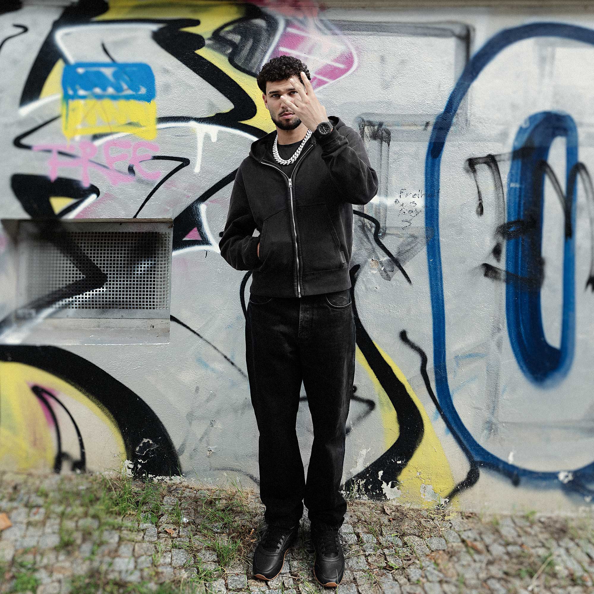 Der Rapper Lucio101 in schwarzer Kleidung mit einer massiven Silberkette und der G-SHOCK "Luxe black" Armbanduhr steht vor einer mit Graffiti besprühten Hauswand.