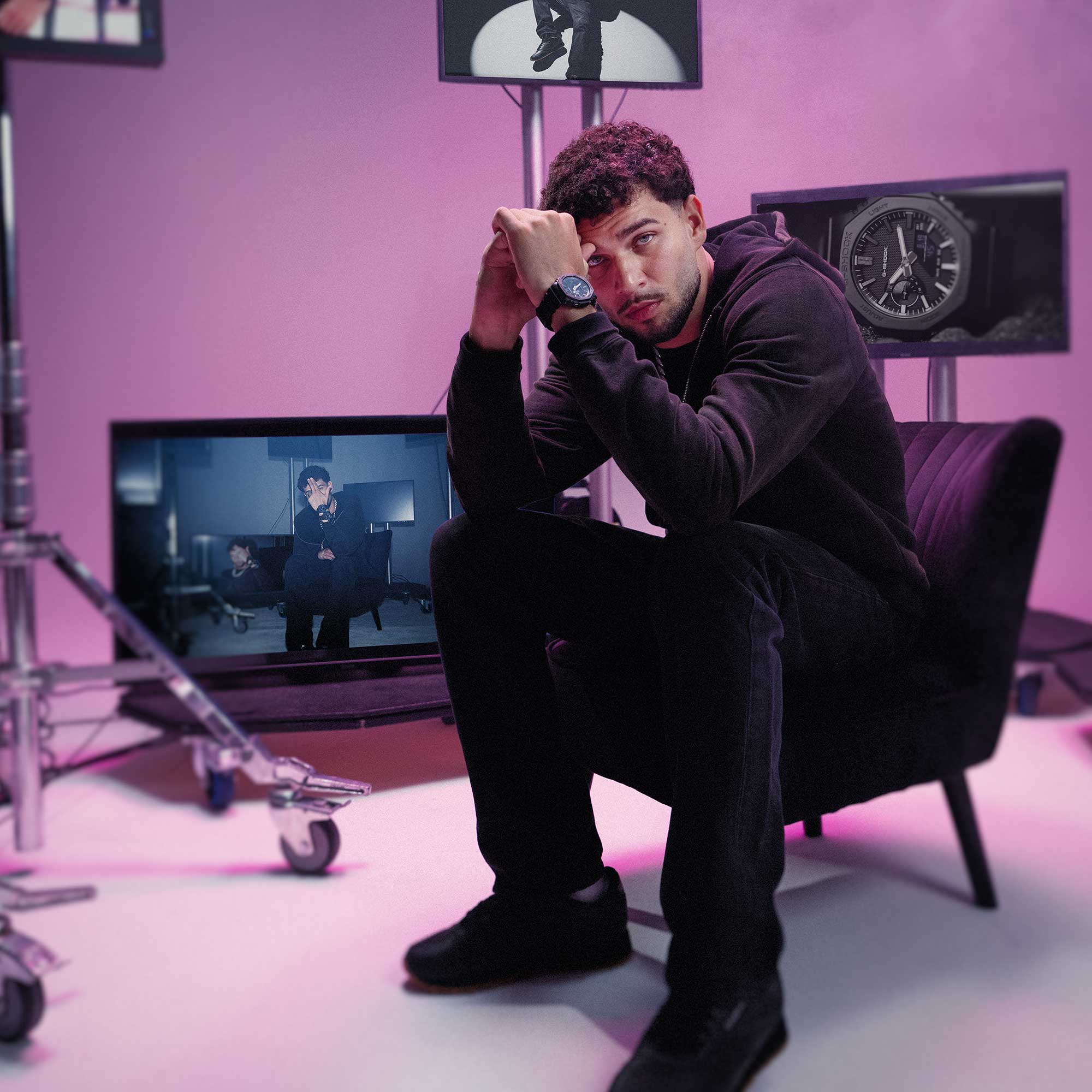 Lucio101 sitzt auf einem schwarzen Sessel in einem pinken Studio mit schwarzer Kleidung, schwarzen Sneakern und der schwarzen Luxe Black Armbanduhr. Im Hintergrund sind man auf mehreren Bildschirmen die Uhr GA-2100LXB.