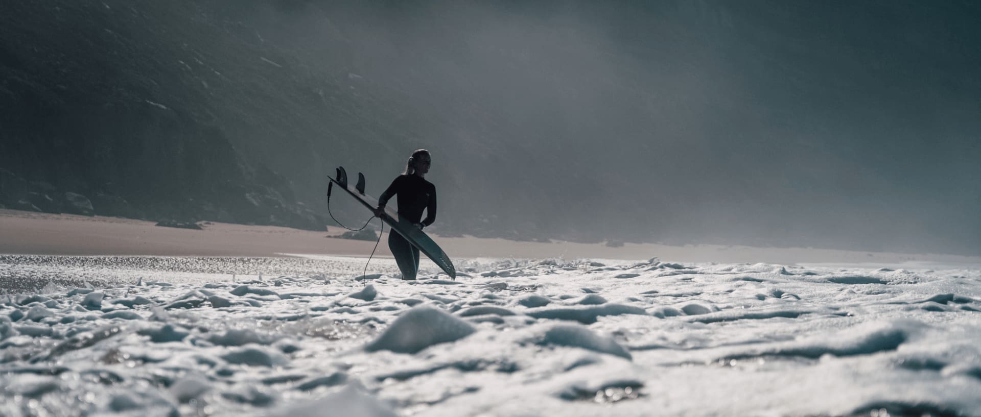 Christina Gindl steht mit Surfboard im schäumigen Meer und Weißwasser