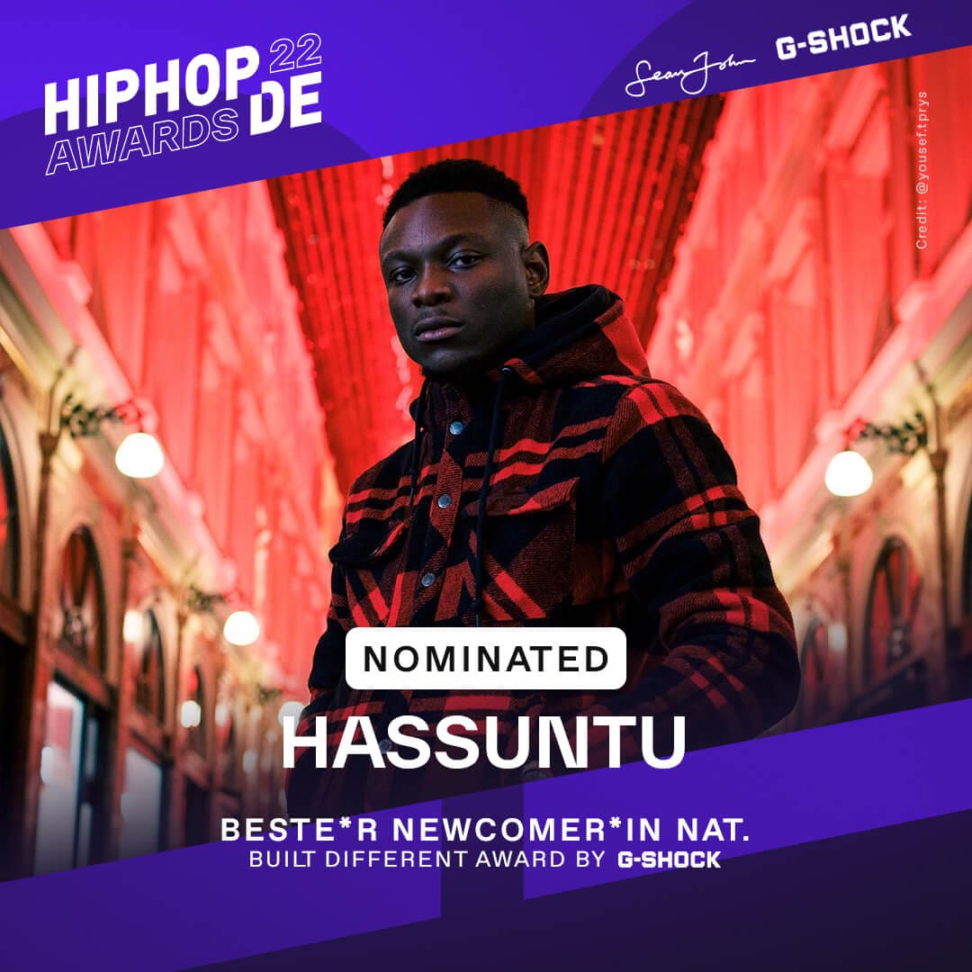 Rapper Hassuntu trägt eine rot schwarz karrierte Jacke, im Hintergrund ist eine rot-beleuchtete Einkaufspassage zu sehen. Er war als bester Newcomer national nominiert.