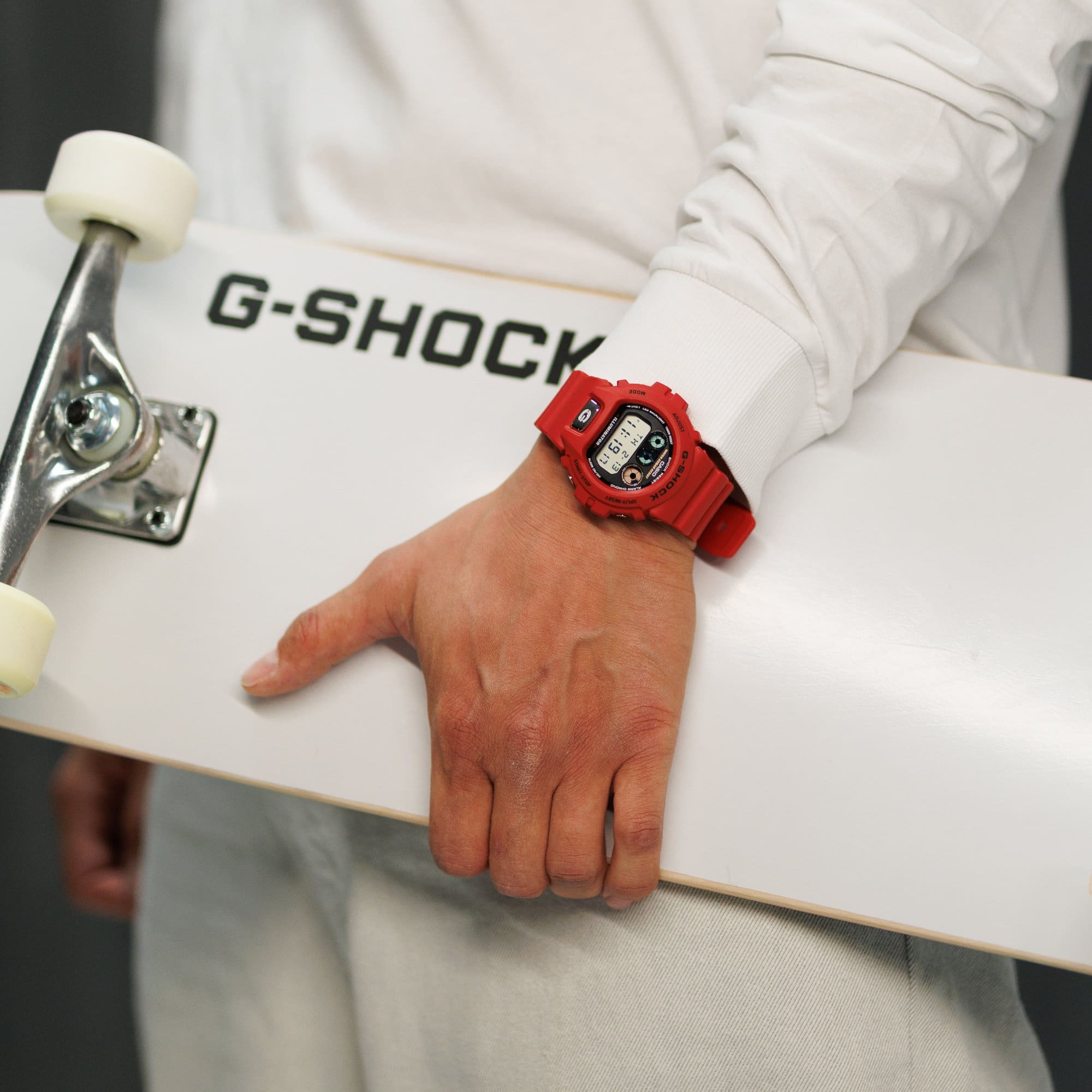 Ein weiß gekleideter Mann trägt ein Skateboard und die rote Digitaluhr "DW-6900TR-4ER" am Handgelenk