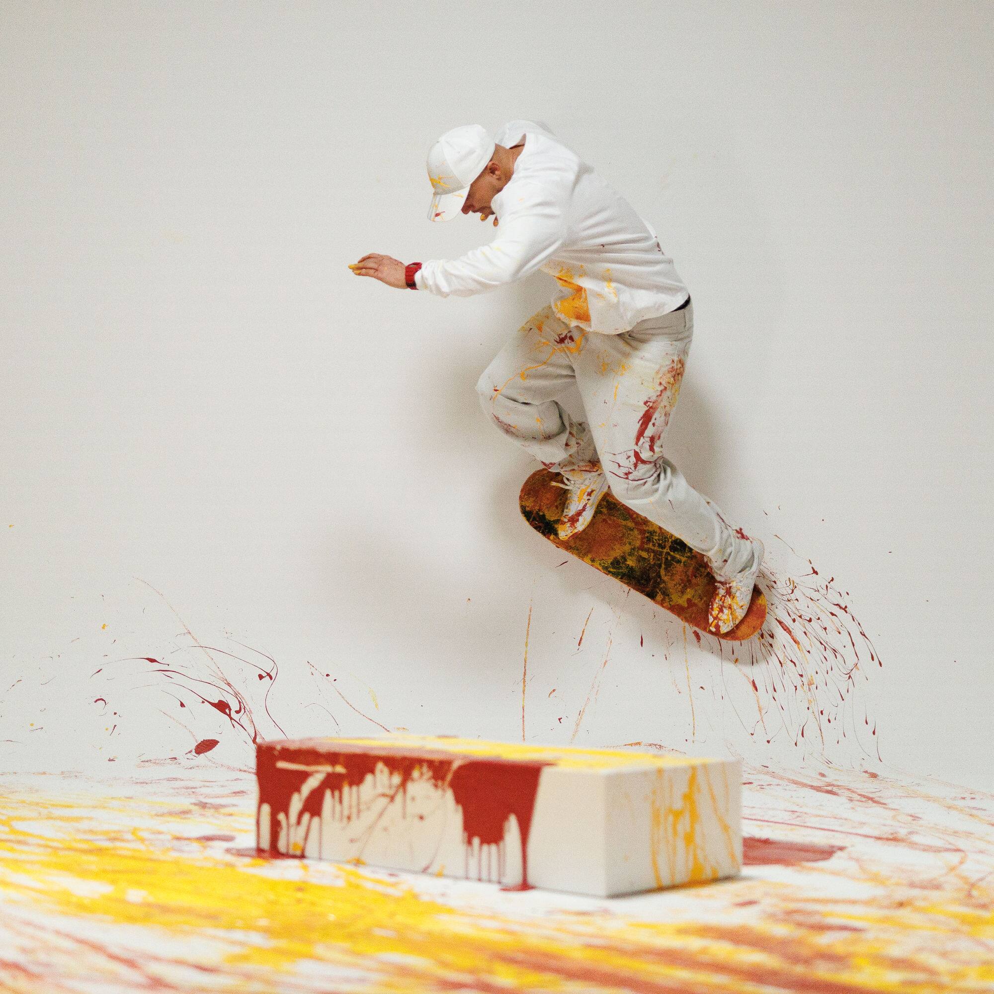 Ein weiß gekleideter Mann mit einer weißen Mütze fährt mit seinem Skateboard durch einen weißen Raum, der mit roter und gelber Farbe besprenkelt ist.