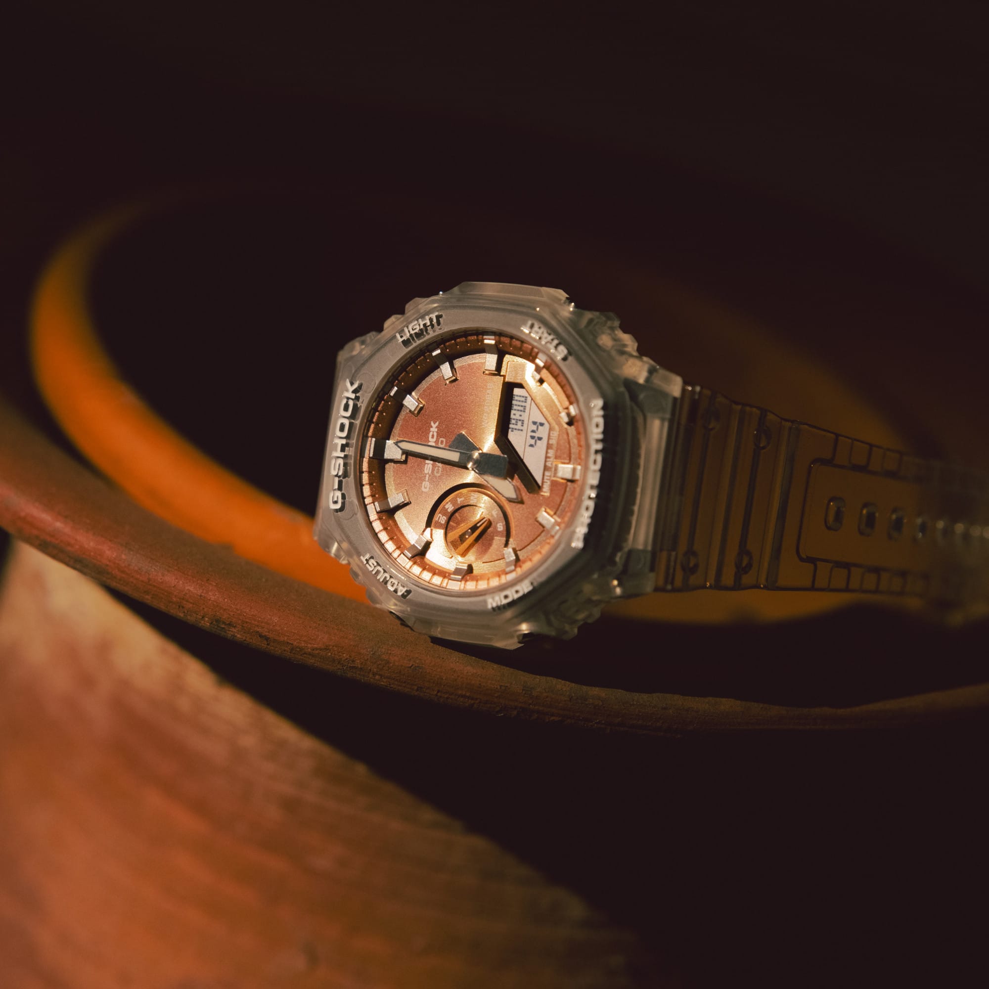 Die GA-2100BM Armbanduhr von G-SHOCK mit transparentem Armband und achteckigem Gehäuse sowie rosé-orangefarbenem Zifferblatt auf einem Tonkrug.