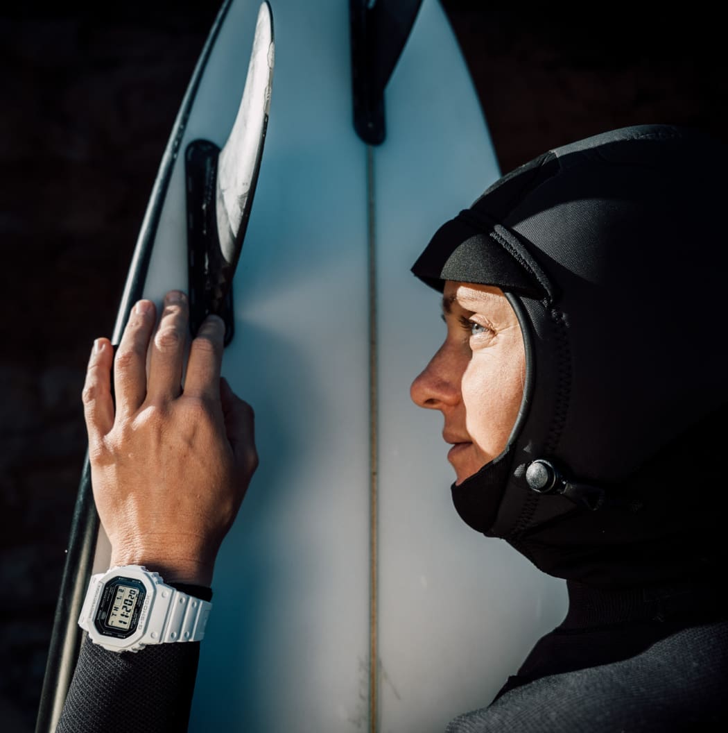 Eine Frau in einem Neoprenanzug trägt die weiße Digitaluhr GW-5000 und hält ein Surfbrett.