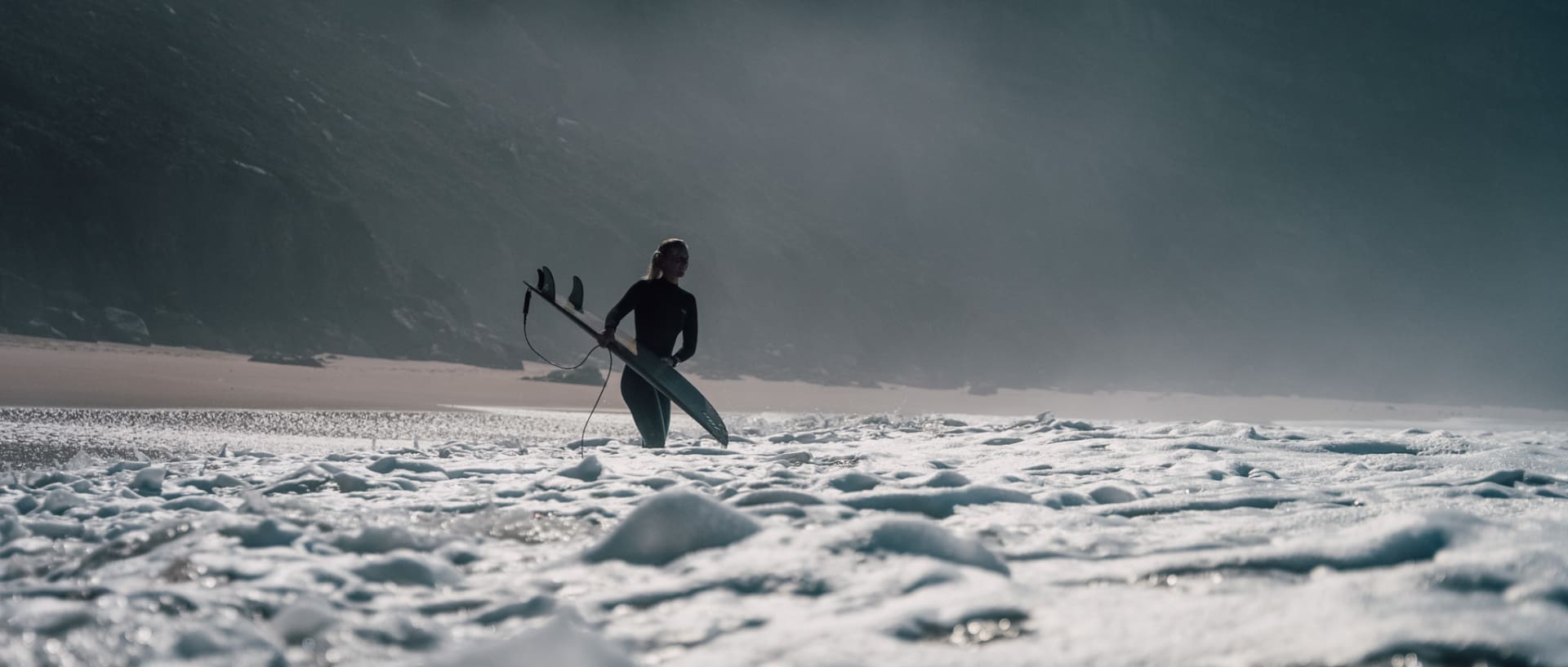 Eine Frau trägt den GW-5000HS und watet mit ihrem Surfbrett in die schäumende See.