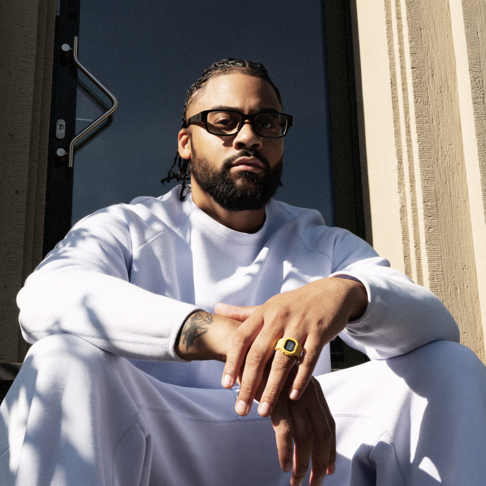Ein ganz in Weiß gekleideter Mann sitzt auf einer Treppe und hält die Hand vor sich. Er trägt die gelbe digitale Ringuhr.
