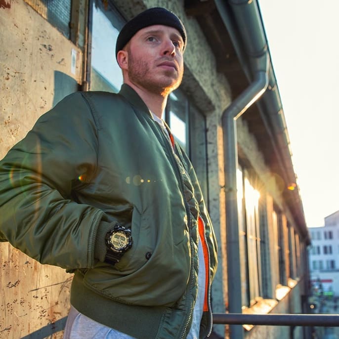 Una persona con chaqueta verde oliva y gorra negra delante de un edificio, lleva el reloj GA-710GB-1ALTD de G-SHOCK en la muñeca.