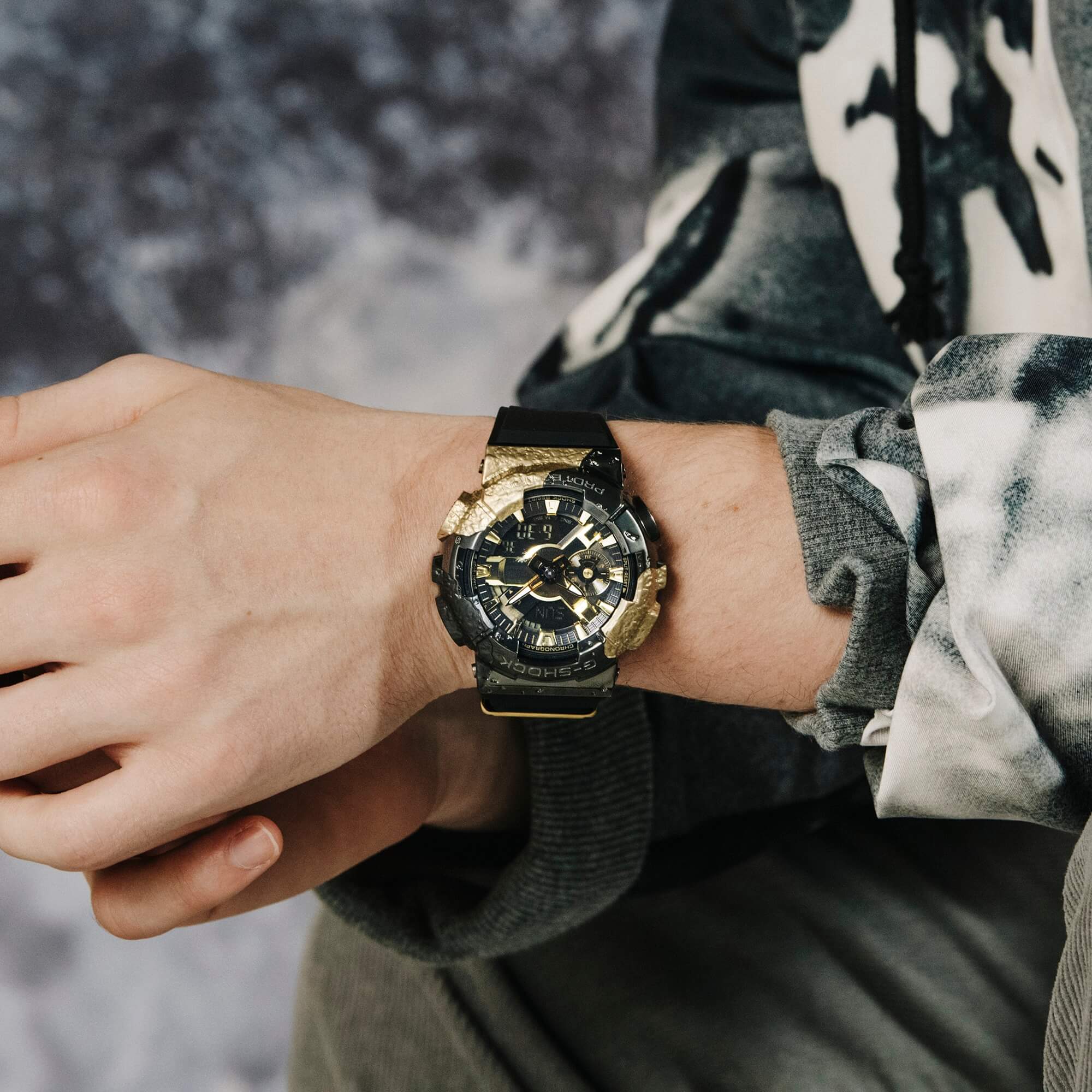 Una persona apoya los brazos sobre las rodillas, el centro de atención es la muñeca en la que se encuentra el reloj de pulsera G-SHOCK GM-114GEM-1A9 con caja de oro macizo y motivos negros y pantalla analógica.