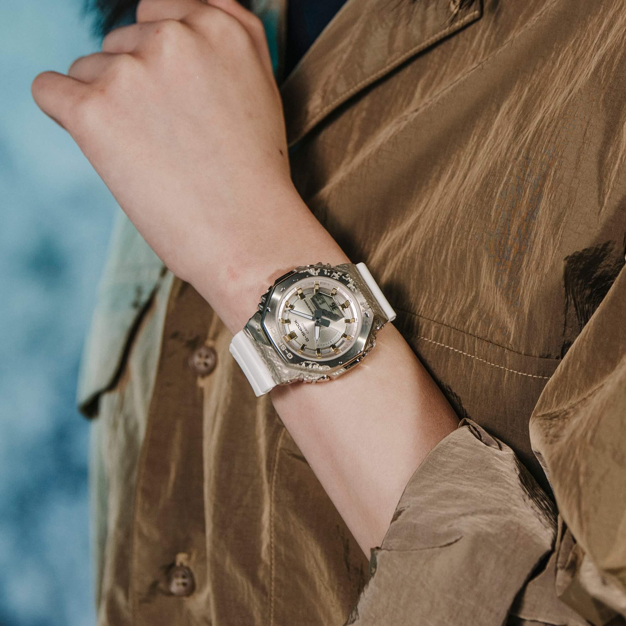 Una mujer viste una blusa marrón y se agarra la muñeca, llevando el reloj de pulsera analógico G-SHOCK GM-S2140GEM-9A con correa blanca y bisel dorado.