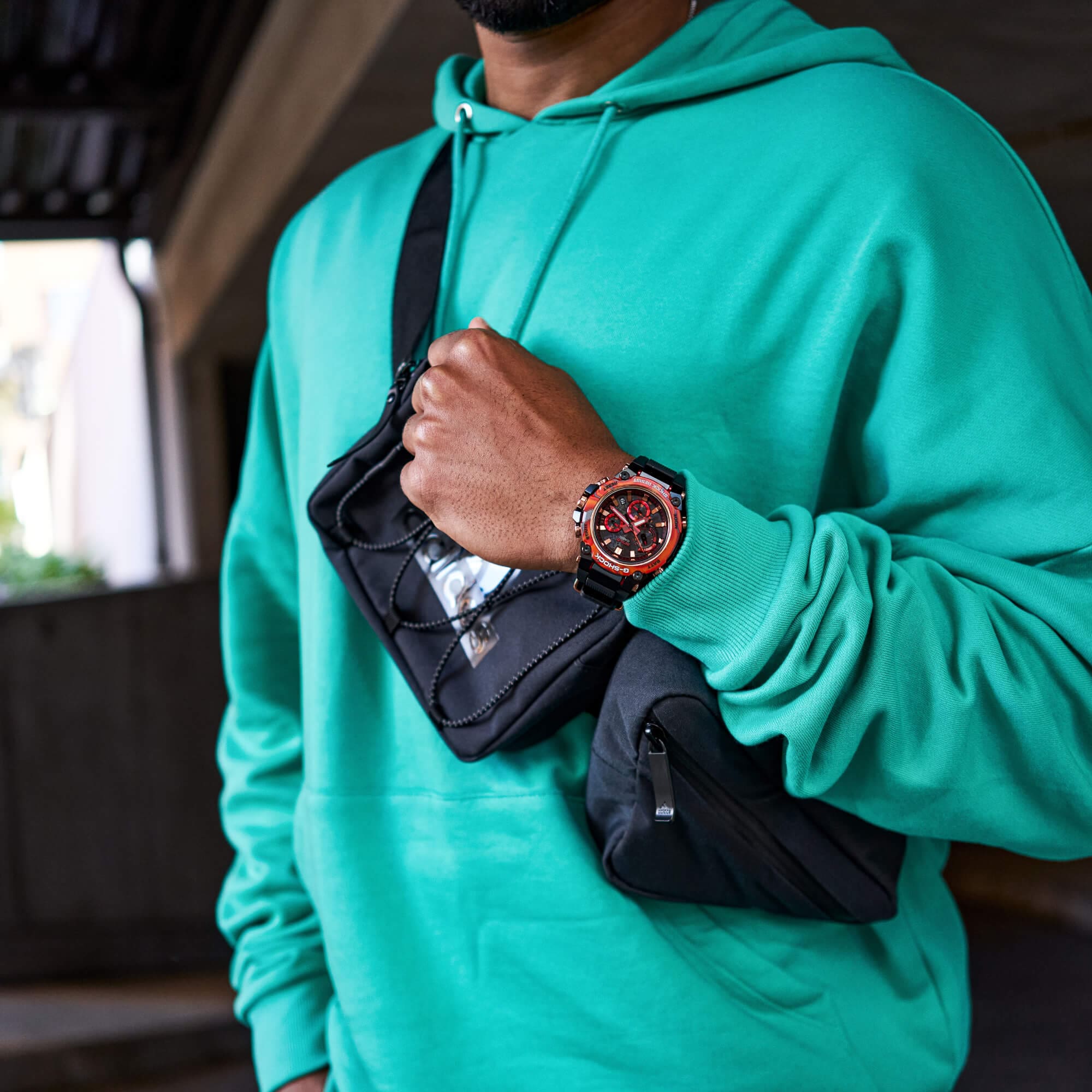 El cronógrafo «MTG-B3000FR-1A» con bisel estampado en rojo y naranja en la muñeca de un hombre que lleva una sudadera con capucha turquesa y un bolso negro cruzado.