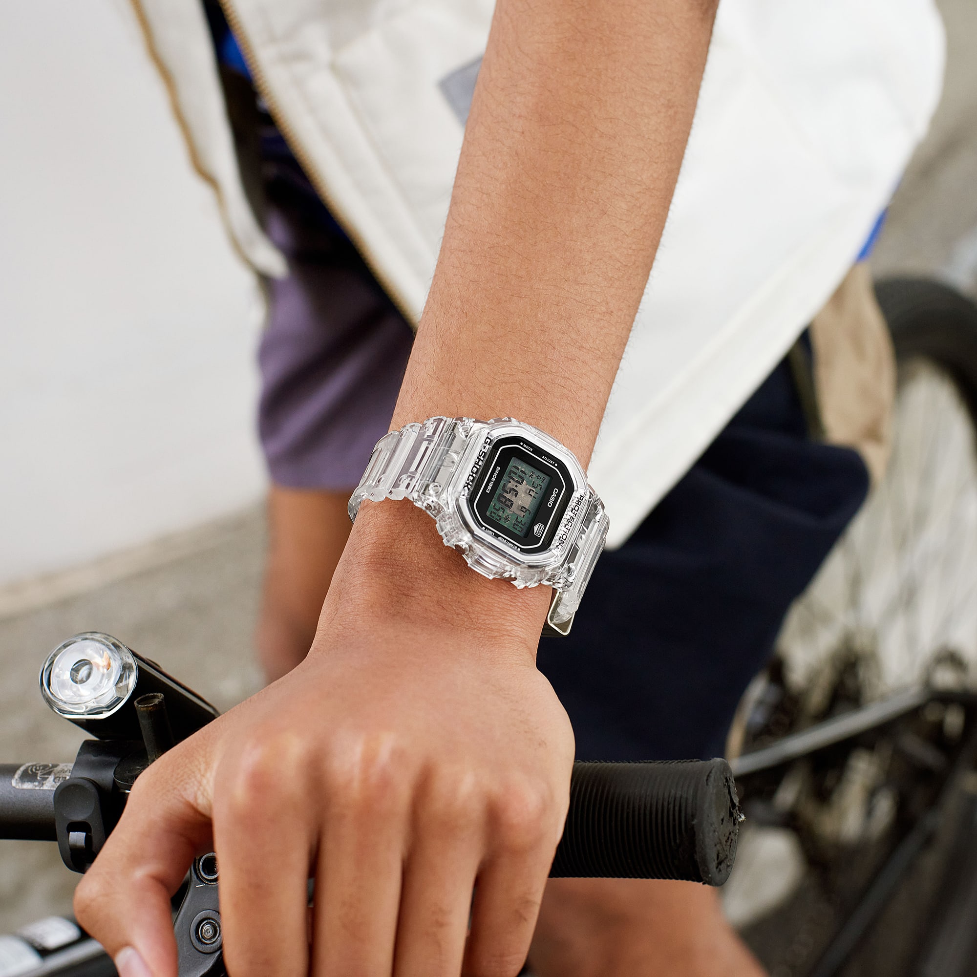 El reloj digital DW-5040RX-7 con brazalete transparente, pantalla rectangular y cierre dorado en la muñeca de un hombre en una moto de carreras.