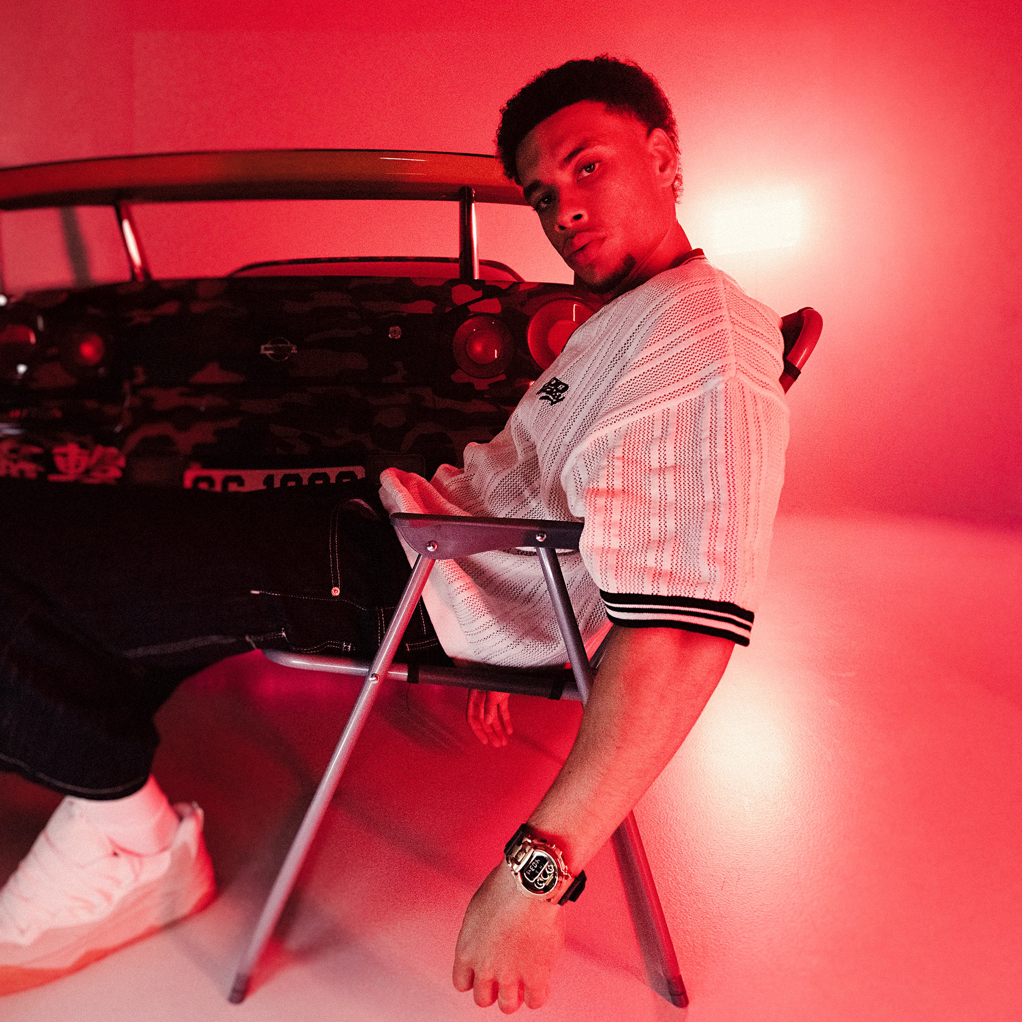 Ein Mann sitzt in rotem Licht in einem Studio auf einem Stuhl vor dem Nissan GT-R in camouflage Beklebung. Er trägt ein weißes Shirt und die GM-6900CMG mit Camouflage-Muster am Handgelenk.