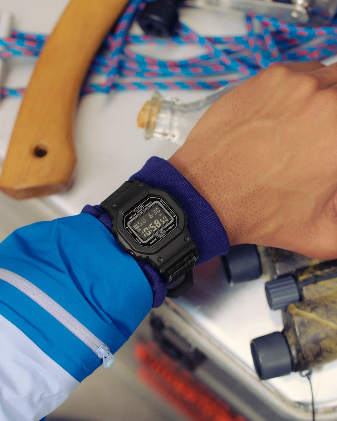 Primer plano del G-SHOCK DW-5600MNC con caja negra en la muñeca, junto a material de montaña como prismáticos y herramientas.