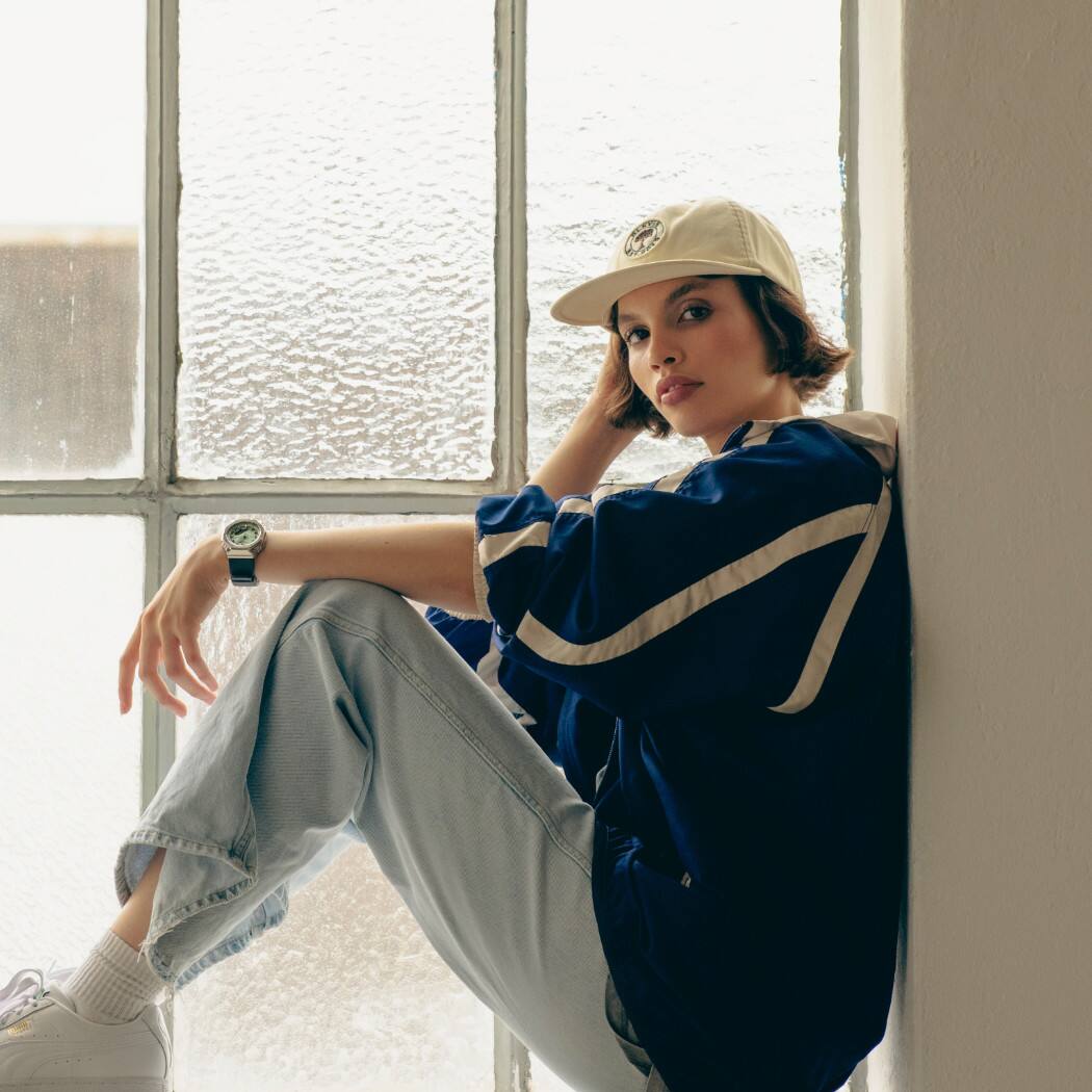 Una mujer morena con gorra sentada en el marco de una ventana, apoyando el brazo sobre la rodilla, lleva el G-SHOCK "GM-S2110-3AER" en la muñeca