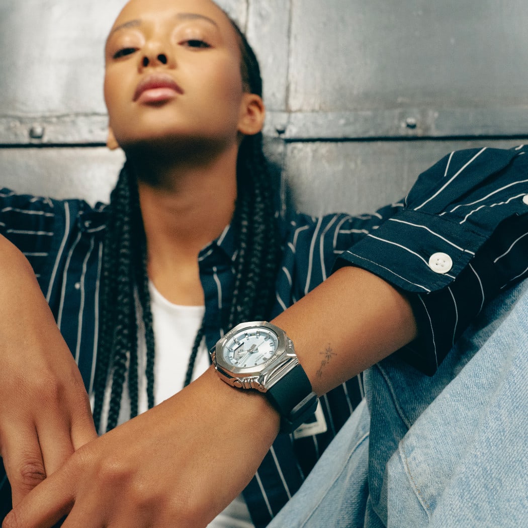 Una mujer está sentada en el suelo, apoyada en una pared metálica y apoyando los codos en las rodillas. El foco de la imagen está en su muñeca, que lleva el reloj de pulsera GM-S2110-2AER.