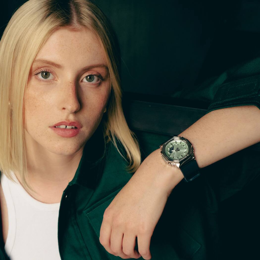 Una mujer lleva el G-SHOCK GM-S2110-3AER con esfera verde en la muñeca y se apoya en una escalera.