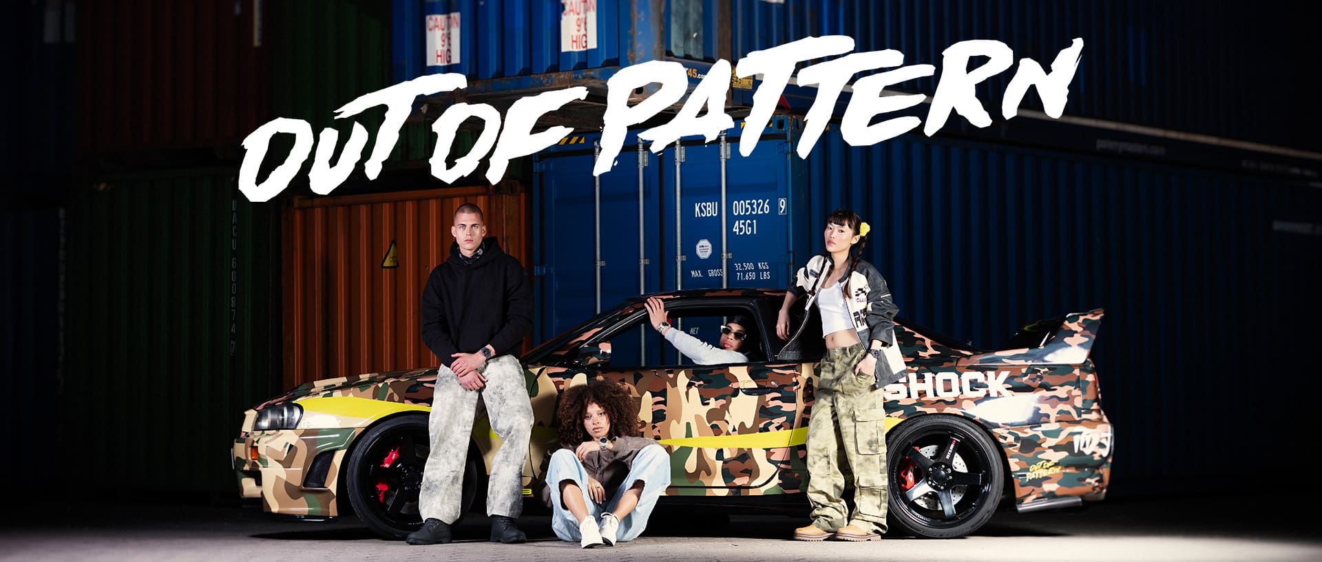 A group of four is sitting in or crouching in front of the Nissan, which is parked in the harbor in front of some containers.