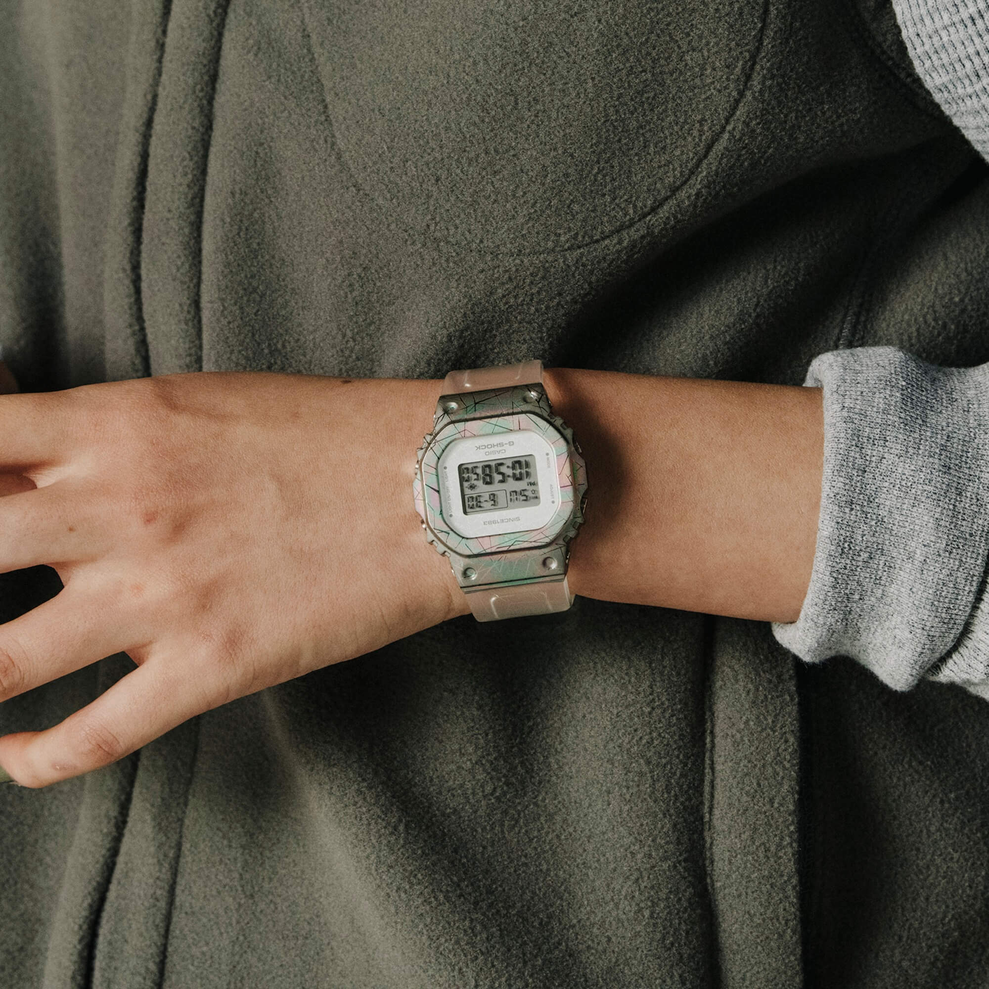 The G-SHOCK GM-S5640GEM-7 wristwatch in white with a silver-pink-turquoise patterned case and digital rectangular display is worn on the wrist.