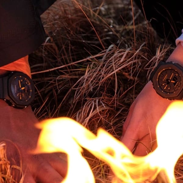 Two people wear variants of the G-SHOCK Utility Black. In front of their hands, a fire can be seen.