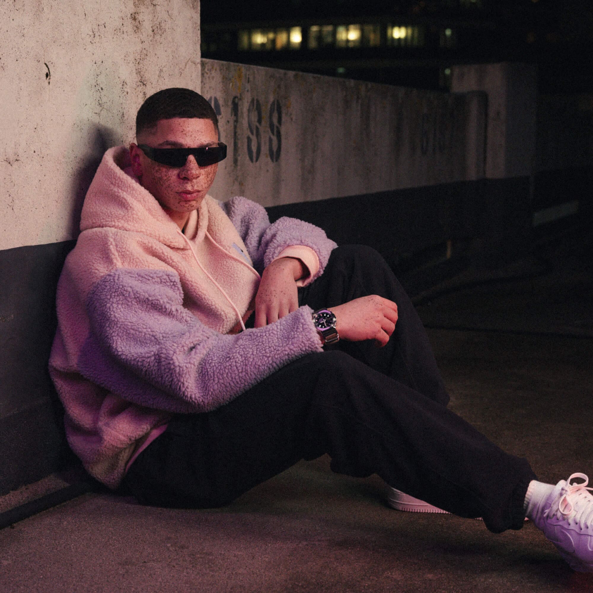 A man sits on a wall in a parking garage wearing sunglasses and the GM-2100CB watch by G-SHOCK
