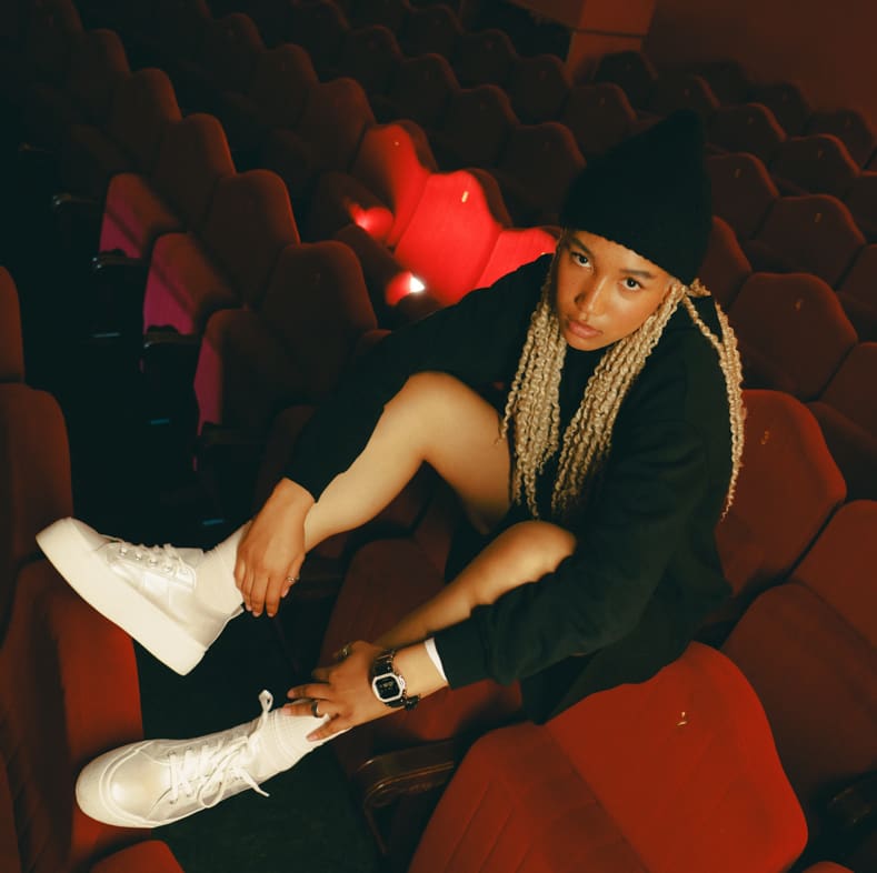 A young woman sits casually in a cinema. She is wearing the GM-5600YM-8ER in silver.