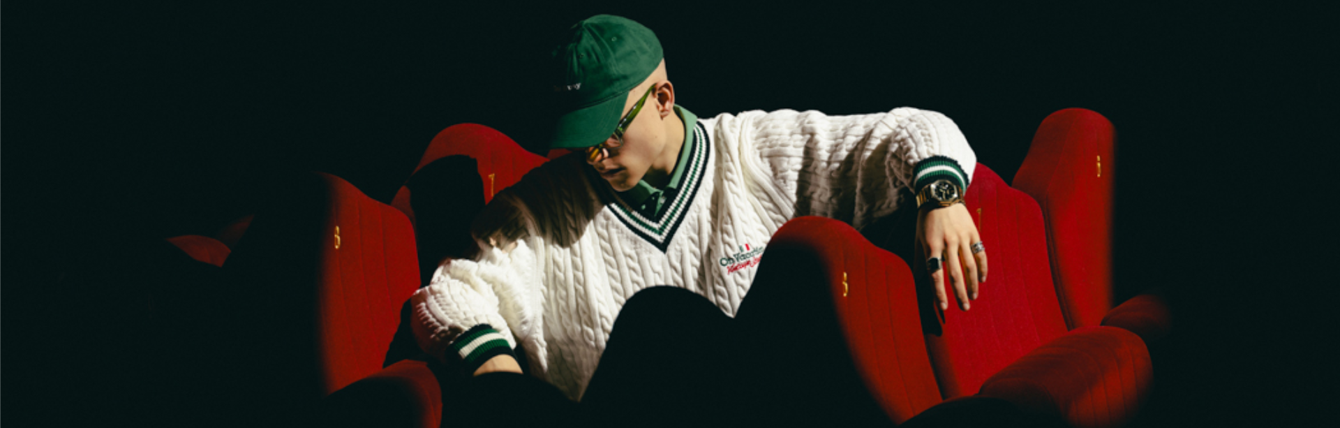 A man in a green cap sits in an empty cinema, his arm casually draped over the backrest, wearing the GM-2100YMG.