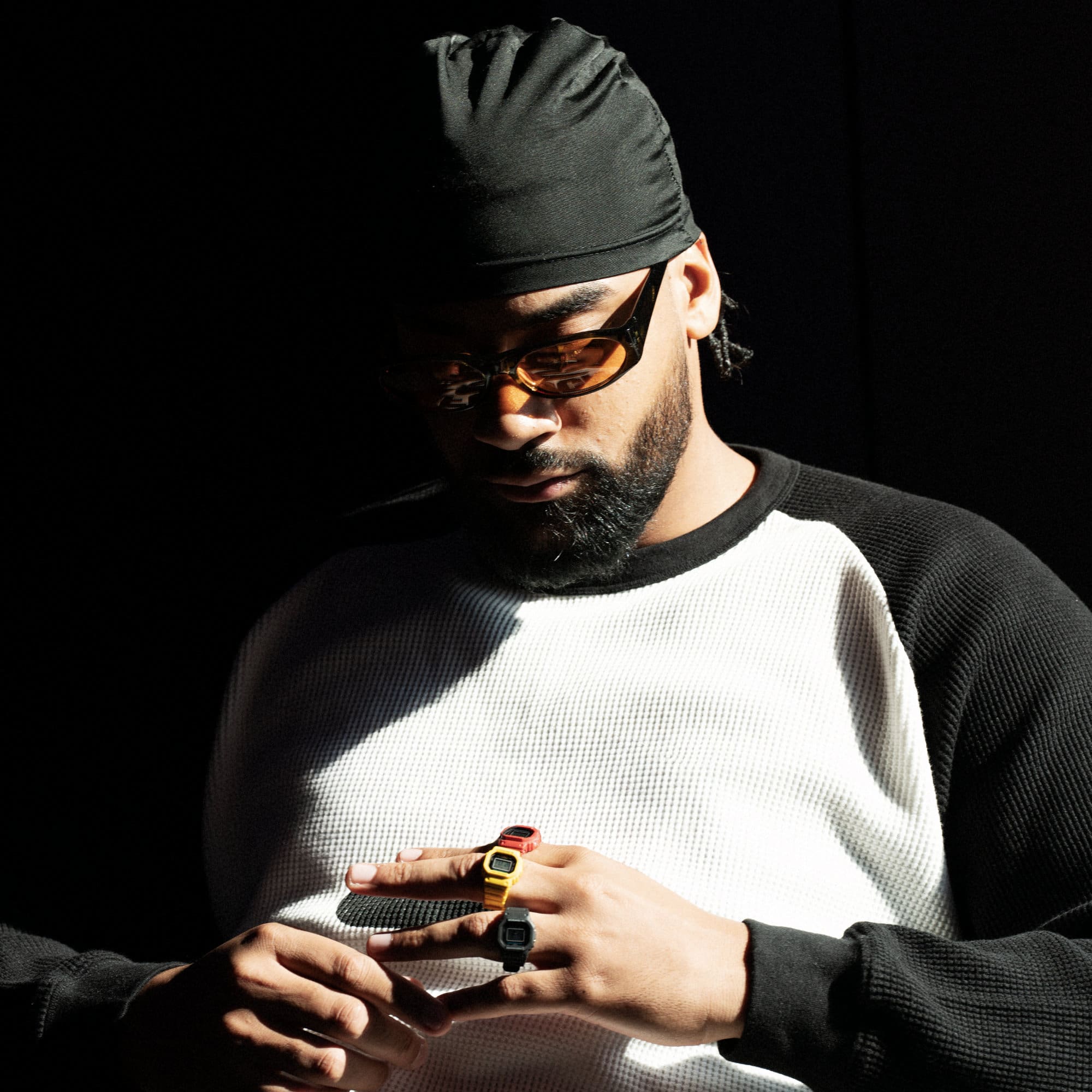  A man with a black cap an orange colored glasses looks down to his hands, he's wearing the three ringwatches in black, yellow und red on his fingers.