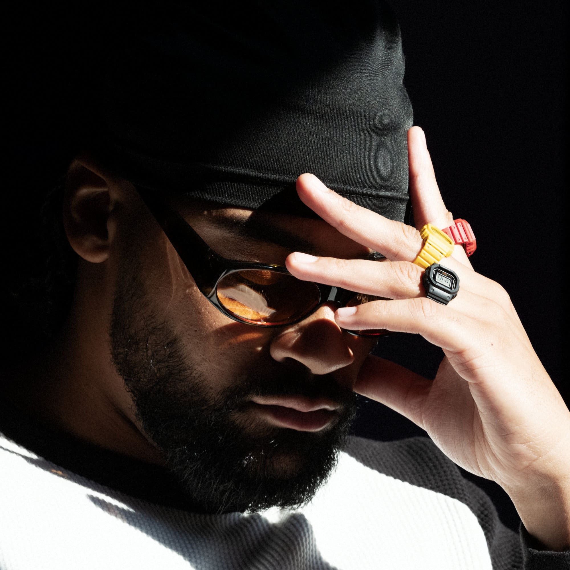 A man holds his hand to his face, he is wearing a black cap, orange glasses and three ringwatches on his fingers.