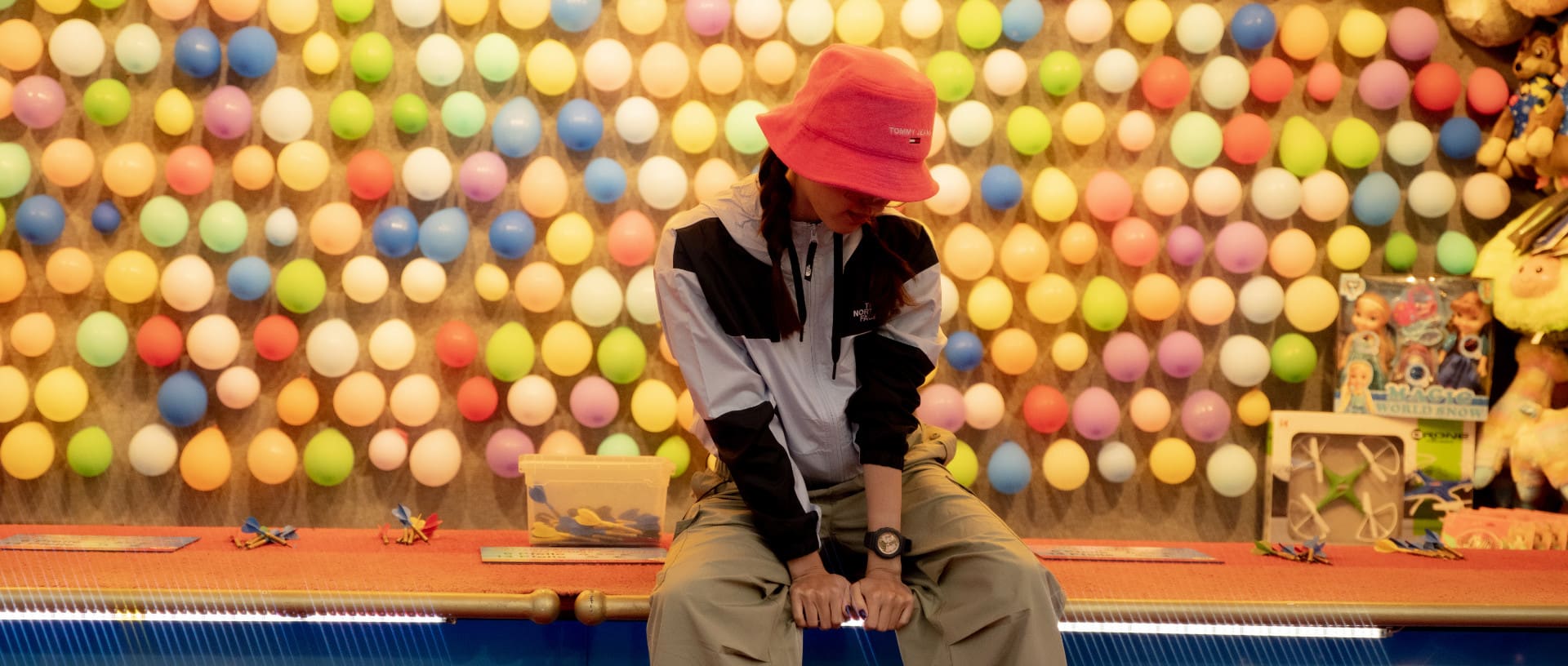 A young woman sits at a colourful fairground stand with baloons wearing the G-SHOCK GMA-P2100SG-1A in black and gold