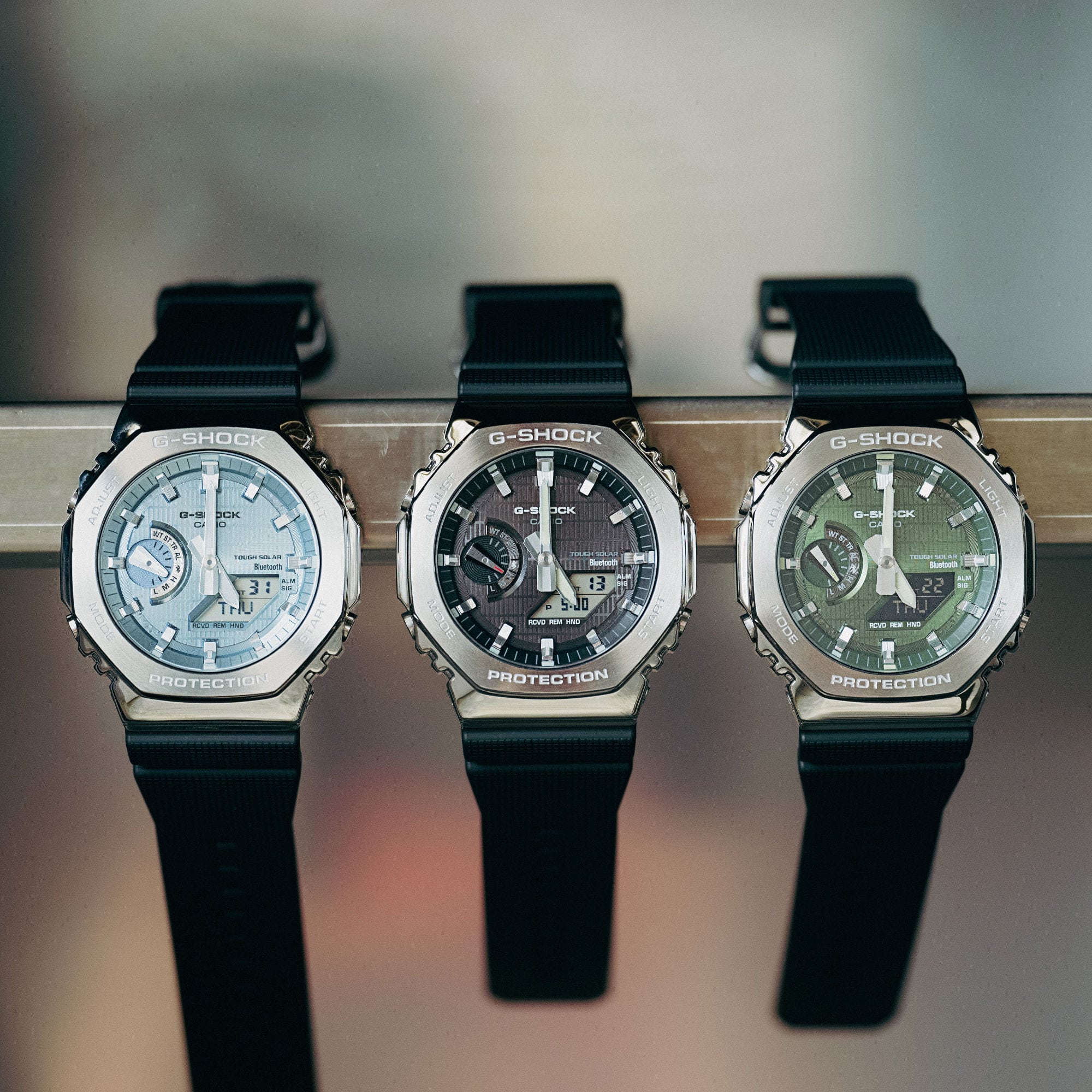 Trois montres G-SHOCK avec bracelets en résine noire et cadran analogique chatoyant en bleu clair, noir et vert suspendues à une barre métallique