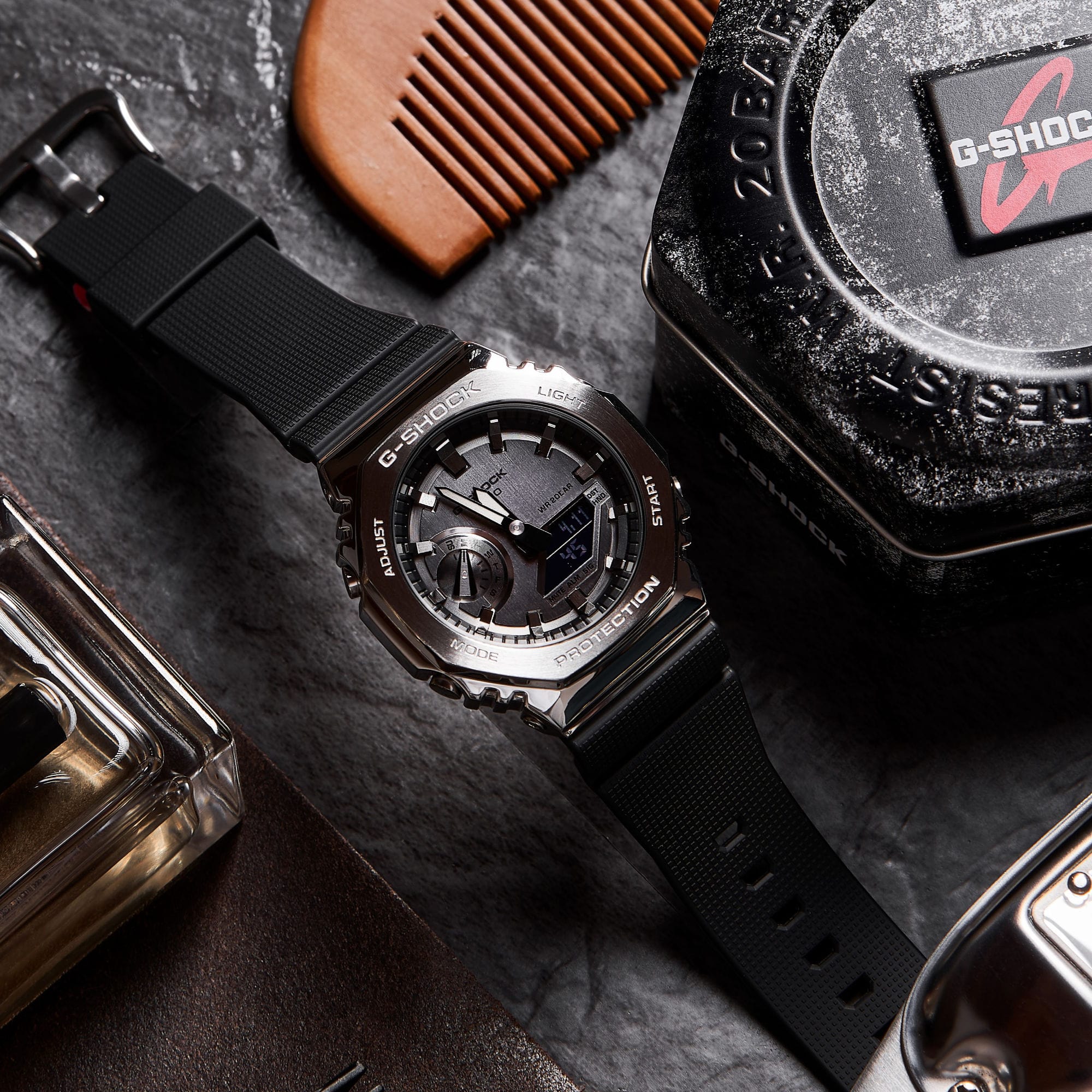 La montre-bracelet GM-2100-1A noire avec cadran analogique noir, bracelet en résine noire et boîtier métallique sur une table, à côté de l'emballage G-SHOCK, d'un peigne, d'un parfum.