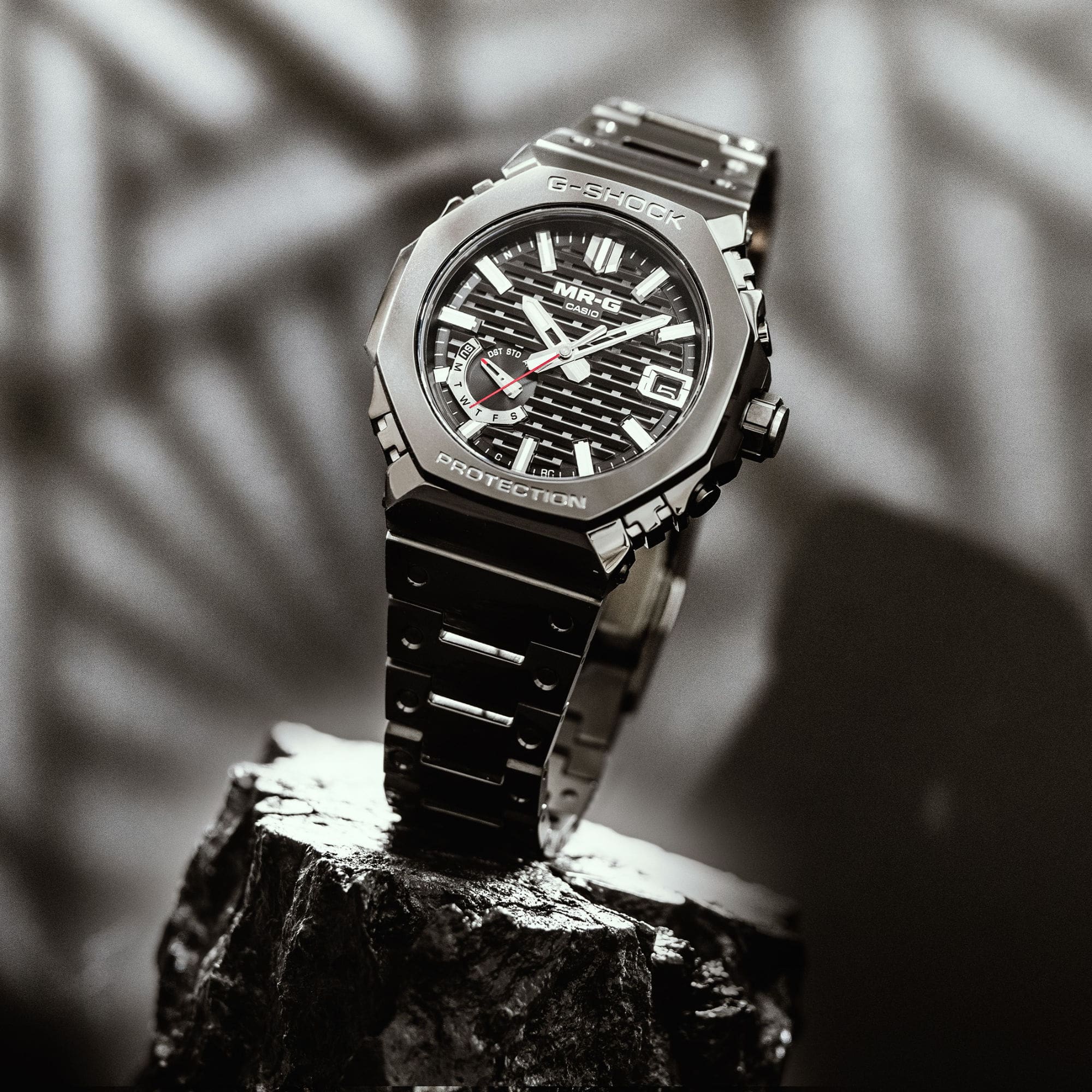 La MRG-B2100B-1A avec bracelet métallique noir et affichage analogique à motifs noirs placé sur une pierre noire