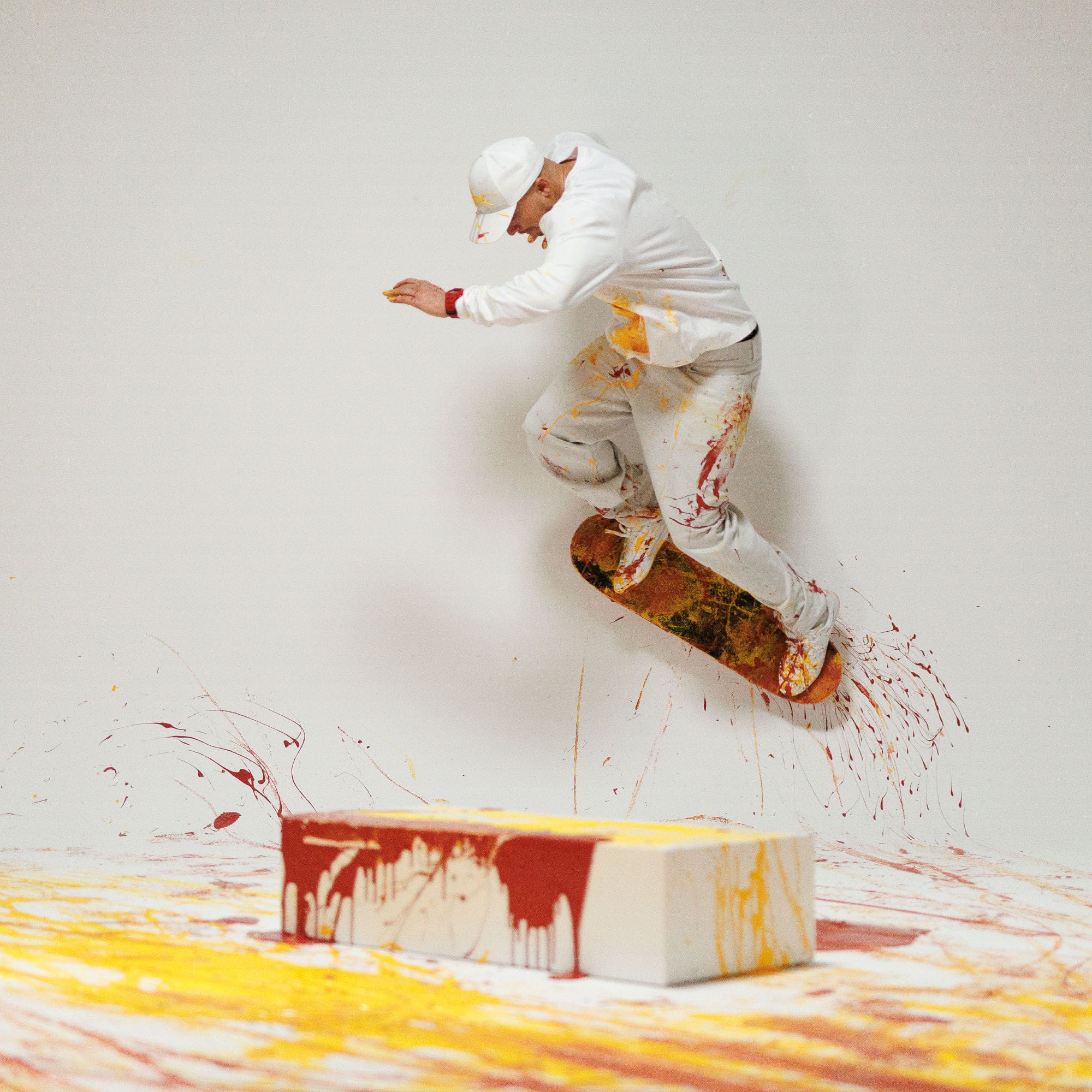 Un homme vêtu de blanc et portant une casquette blanche fait du skateboard dans une pièce blanche parsemée de couleurs rouge et jaune.